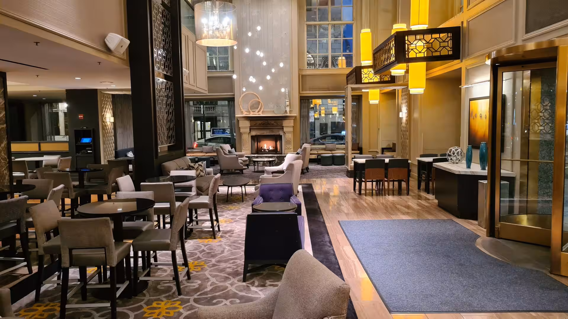 Elegant contemporary hotel lobby featuring lounge seating, tall windows, pendant lighting, a central fireplace, and modern interior design.