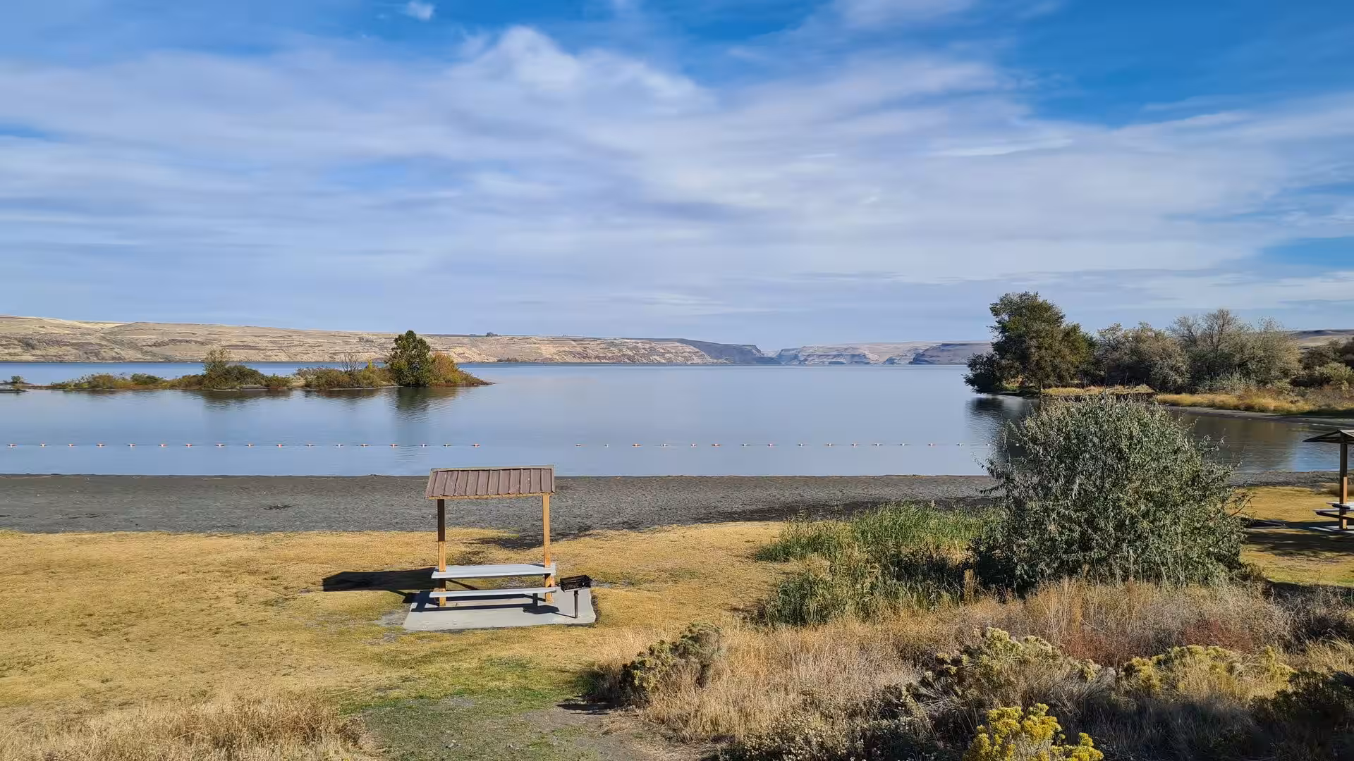 Calm river shoreline with dry grasslands, small islands and distant canyon cliffs under soft clouds