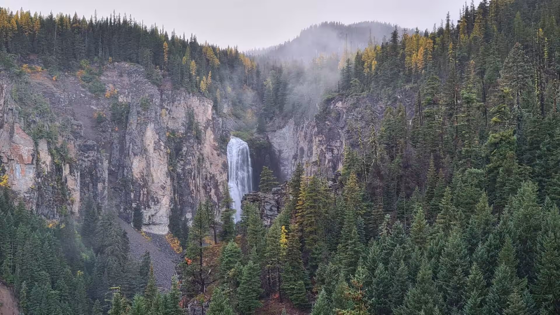 Tall waterfall cascading through a forested canyon surrounded by evergreen trees and drifting mountain mist
