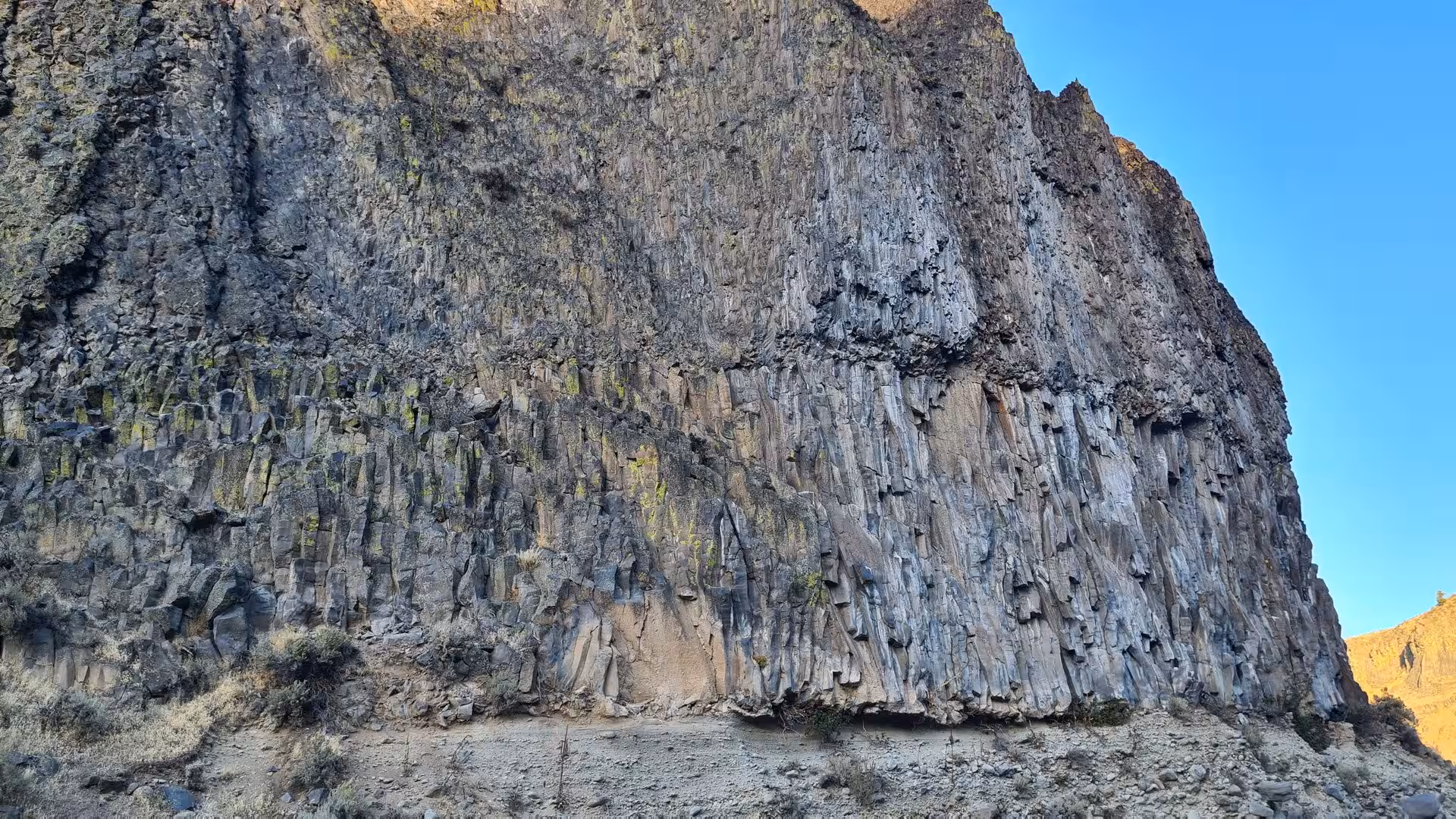 Massive basalt cliff with vertical volcanic columns rising above a canyon floor beneath a clear blue sky