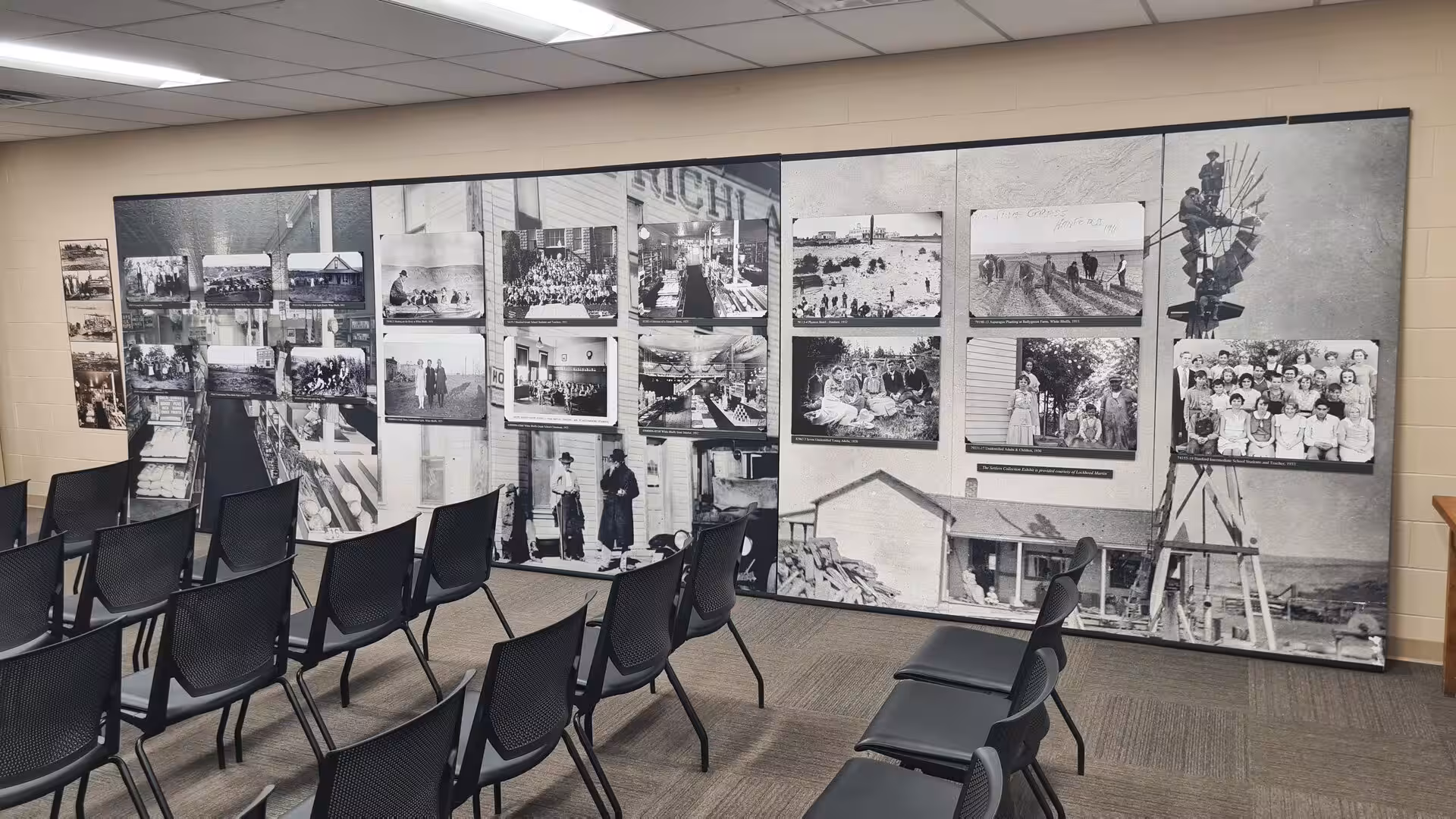 Rows of black and white historical photographs displayed on a large museum wall beside empty chairs