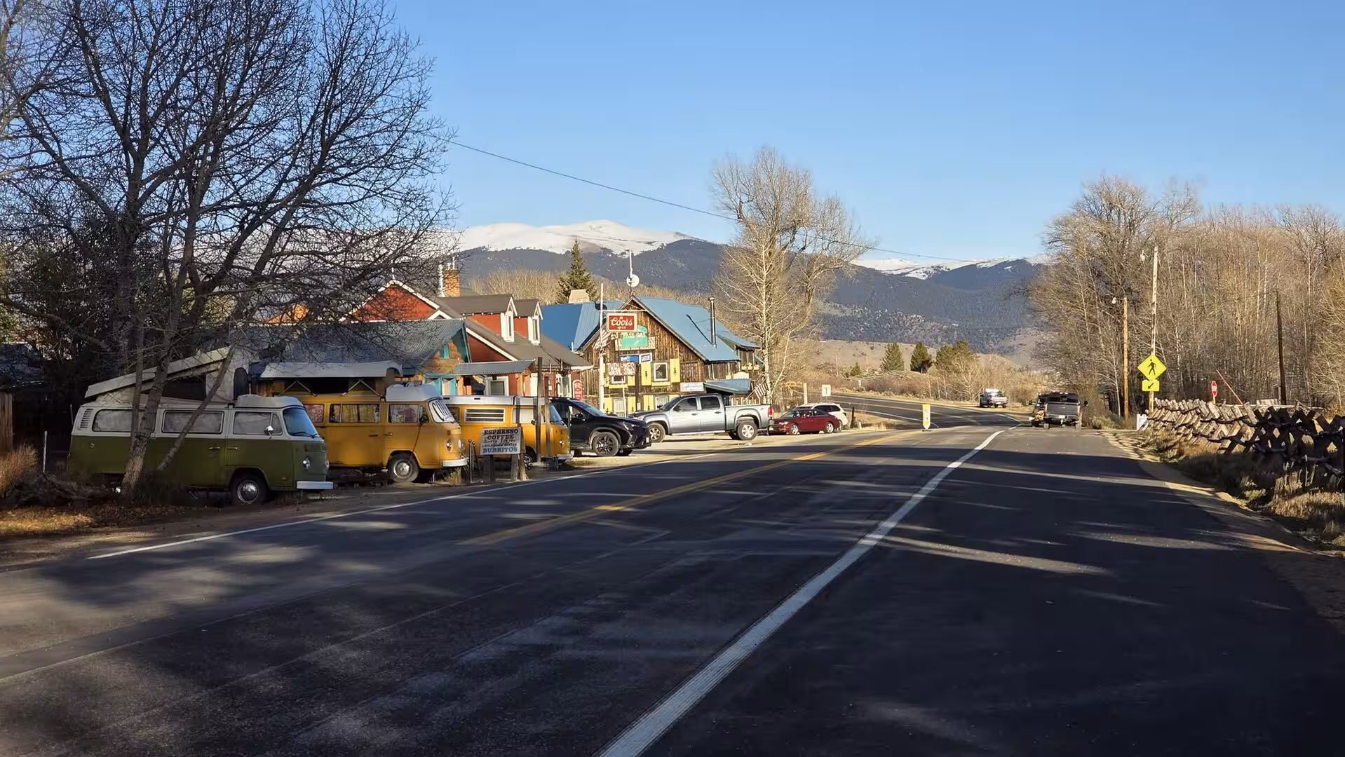 Quiet mountain road passing colorful roadside buildings and vintage vans with snow-covered peaks in the background