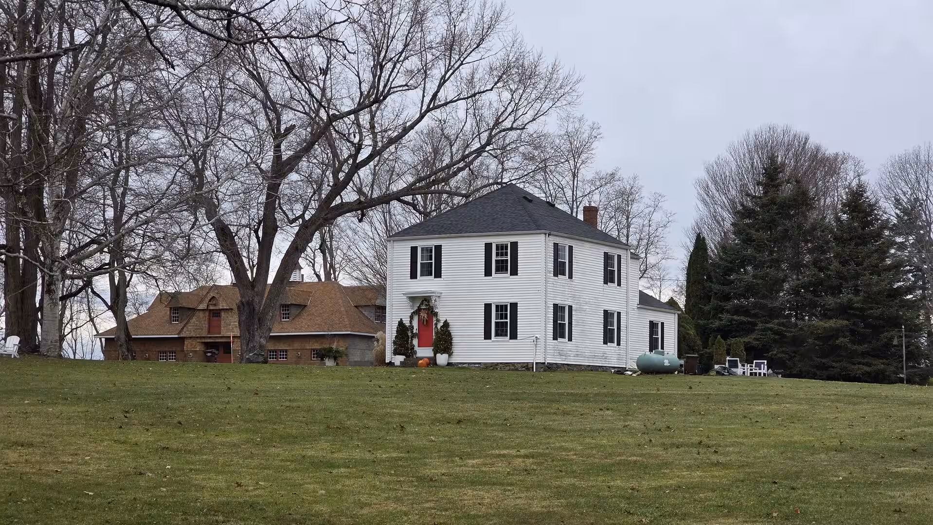 Traditional white farmhouse with red door standing on grassy hill beneath bare winter trees.