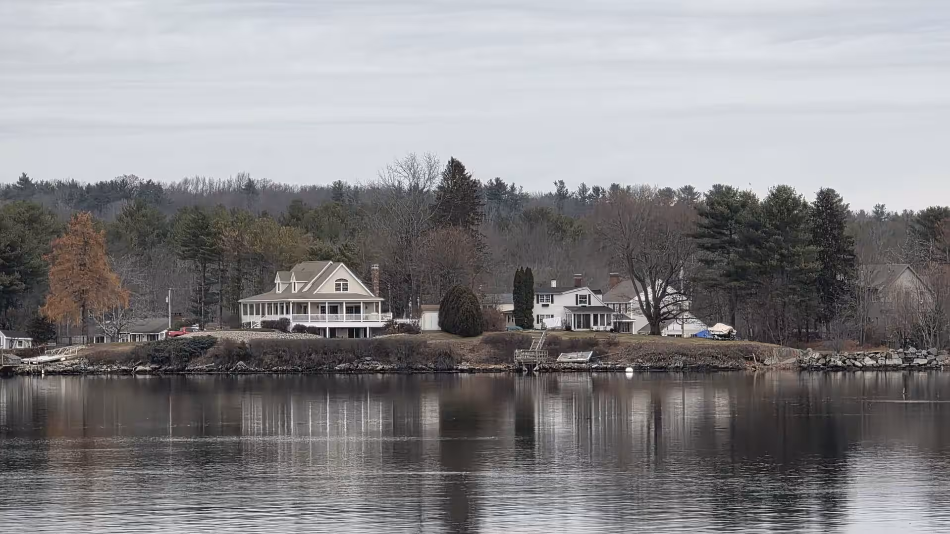Waterfront homes and trees reflected in calm inlet waters beneath gray winter skies.
