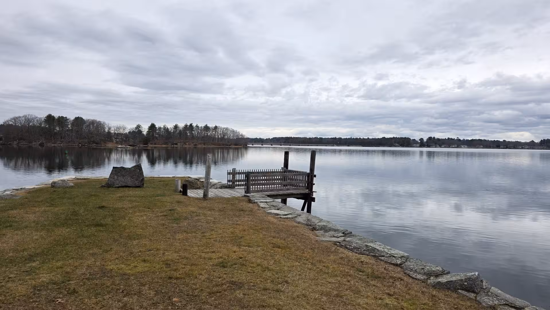 Quiet inlet with small wooden dock, stone shoreline, and calm reflective water under cloudy skies.