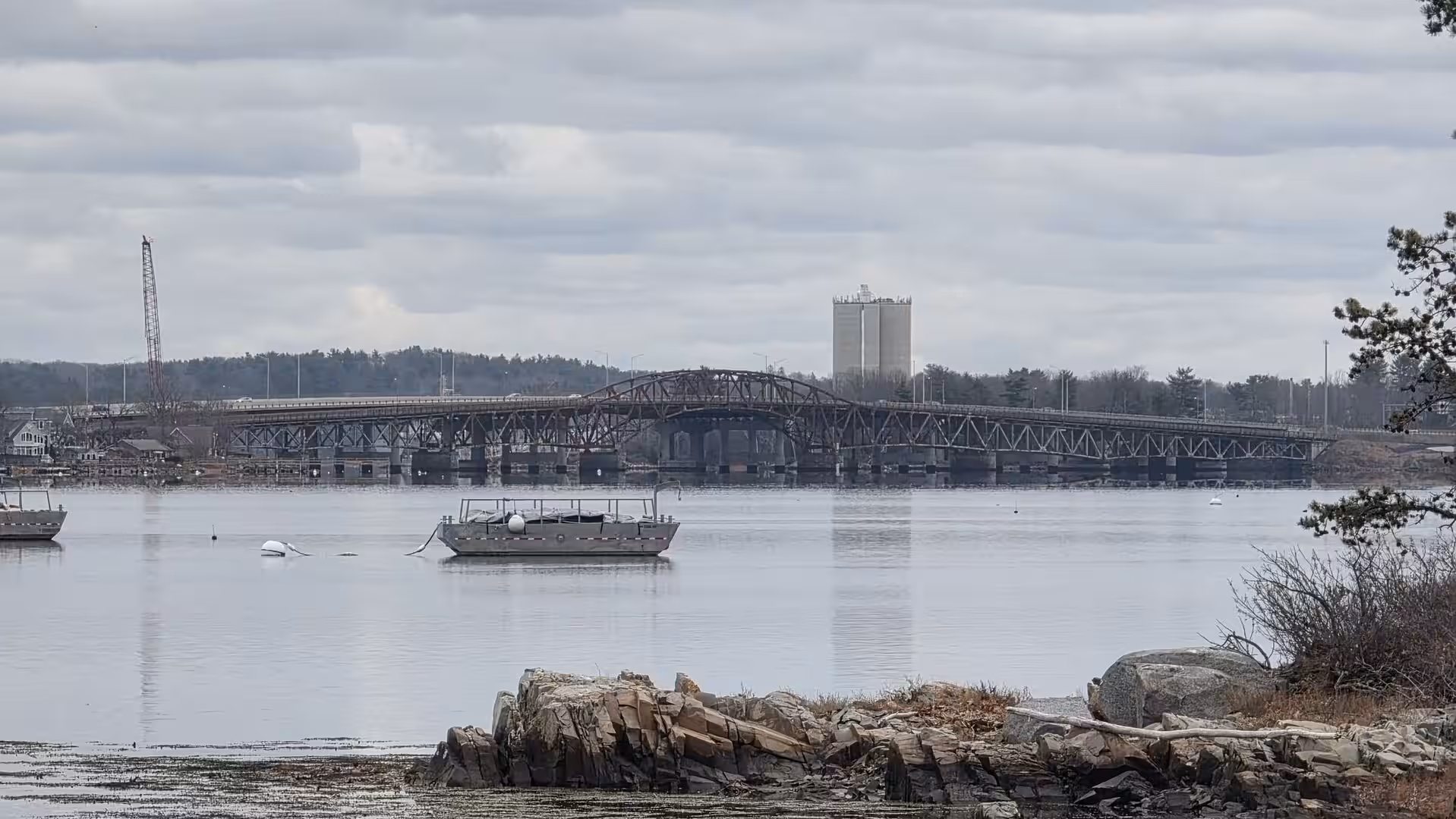 Steel bridge spanning a calm coastal inlet with anchored boat and rocky shoreline in foreground.