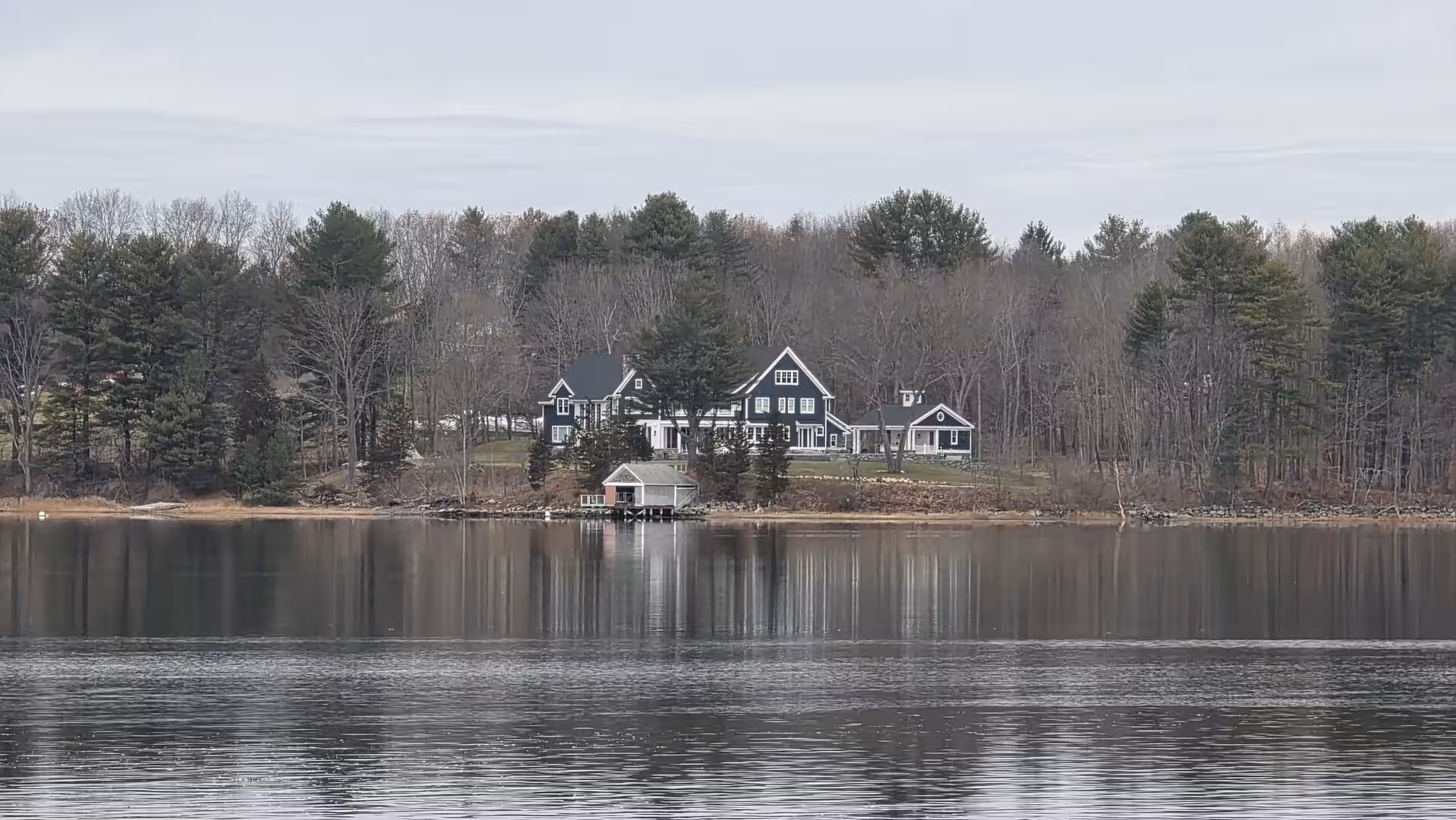 Dark lakeside estate surrounded by pine forest reflected in still water on an overcast day.