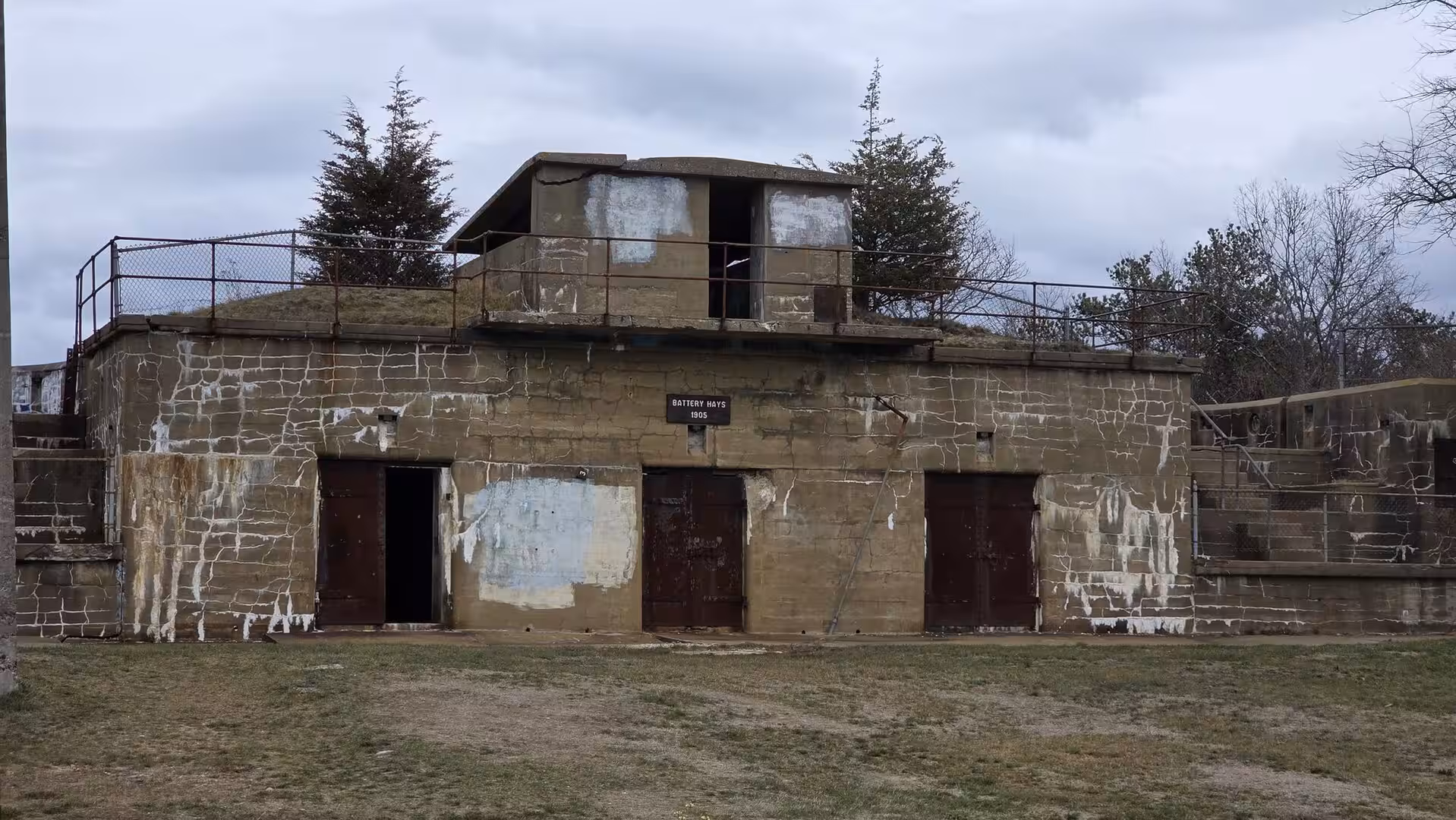 Historic concrete coastal battery bunker with weathered walls, rusted doors, and cloudy skies in New England.