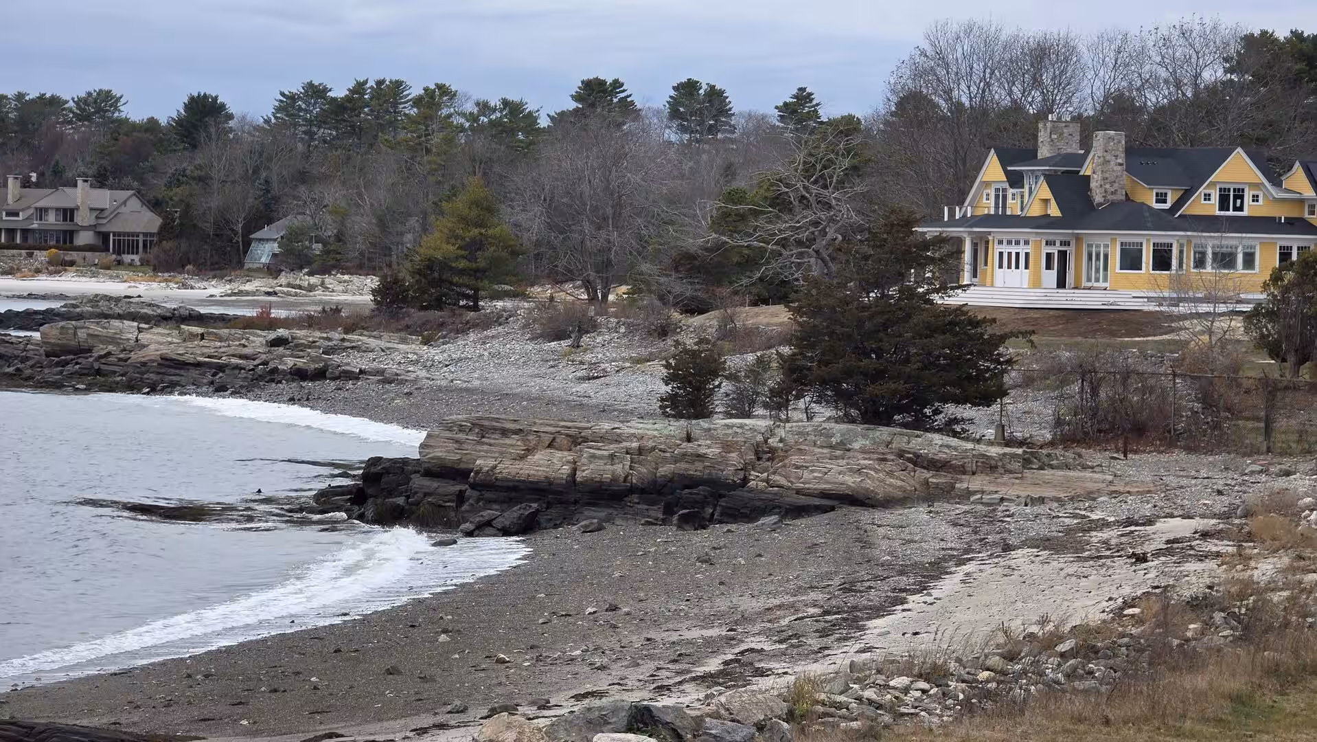 Rocky New England shoreline with a large yellow coastal house overlooking calm ocean water and winter trees.