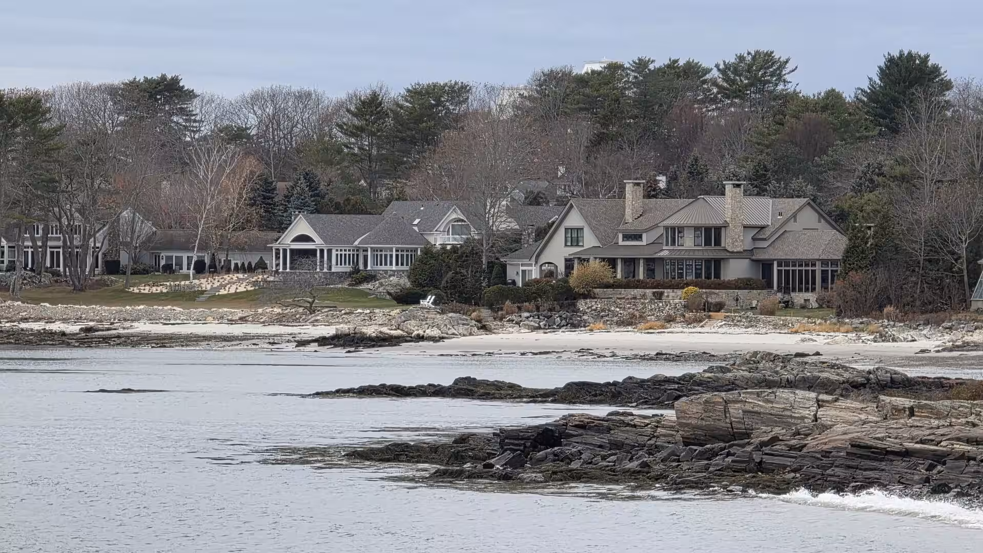 Luxury waterfront homes along a rocky New England shoreline with calm gray ocean water and winter trees.