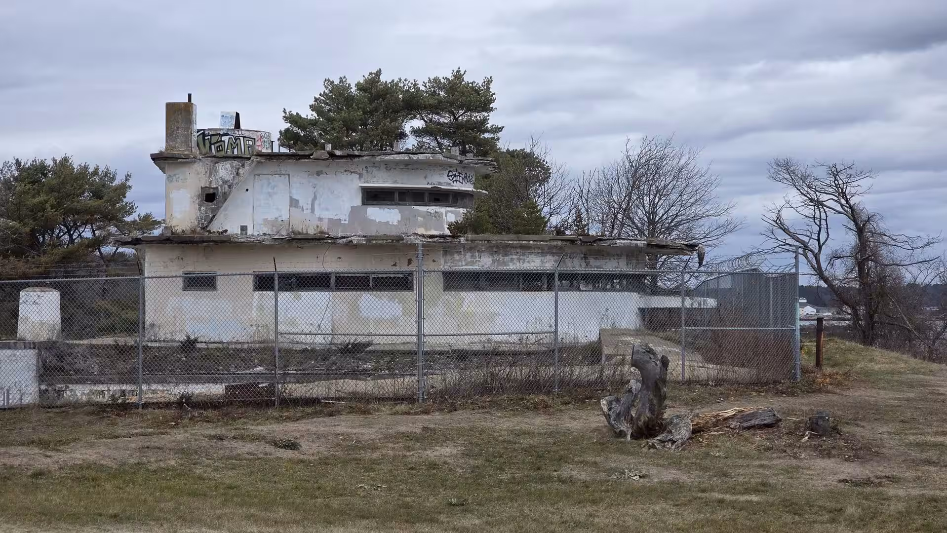 Abandoned coastal concrete bunker surrounded by fencing and trees beneath heavy gray clouds.