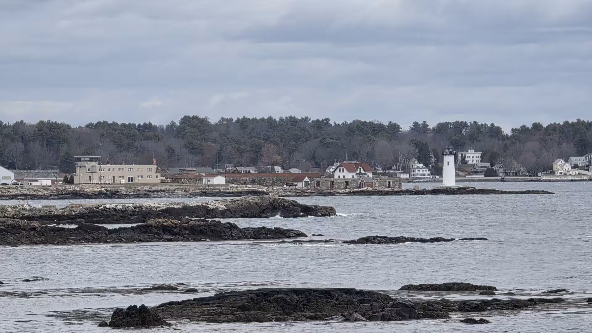 White lighthouse on a rocky New England shoreline with calm coastal waters and dark forest under cloudy skies.