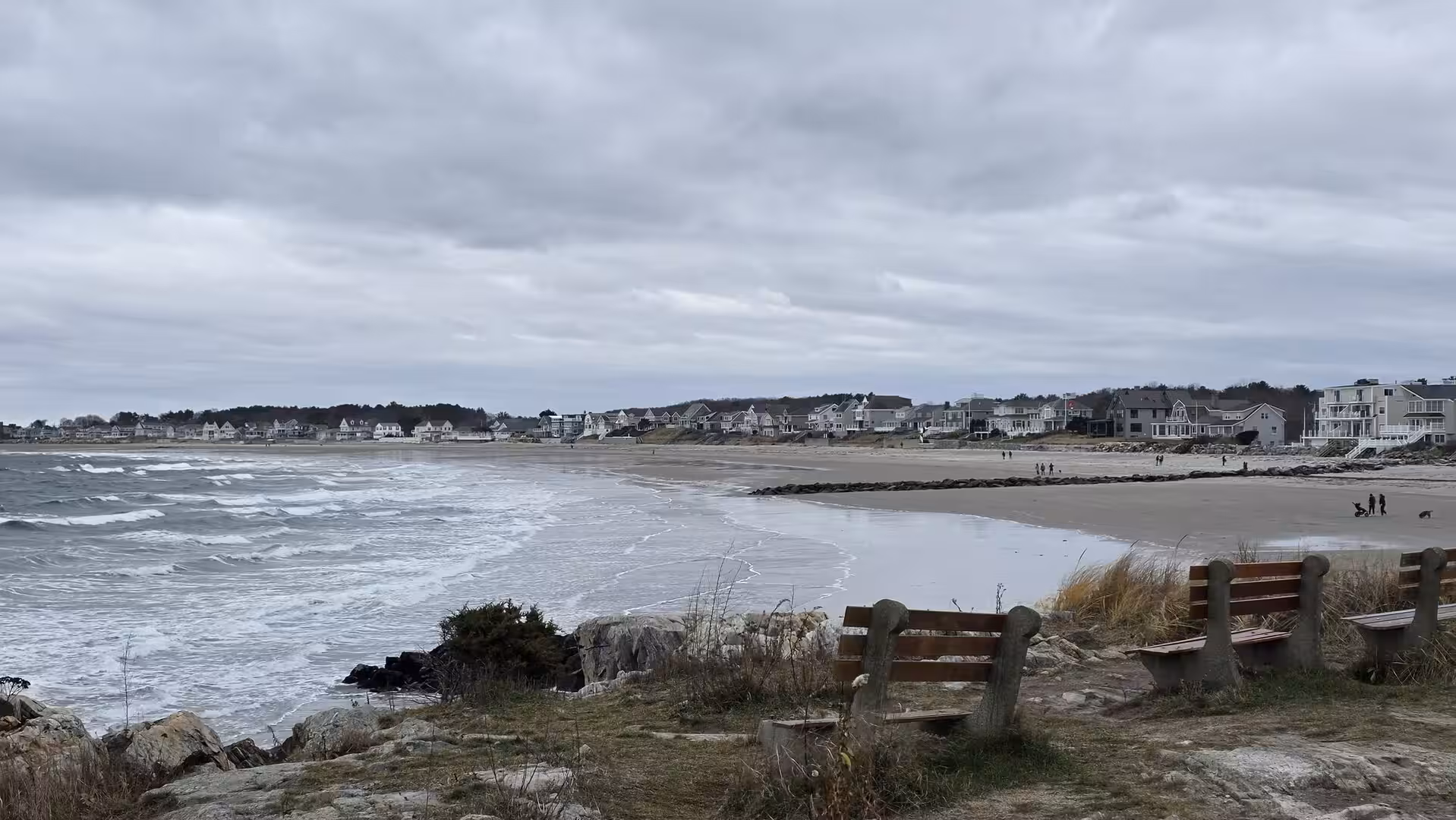 Wide New England beach with rough Atlantic waves, coastal homes, and empty benches beneath dramatic overcast skies.