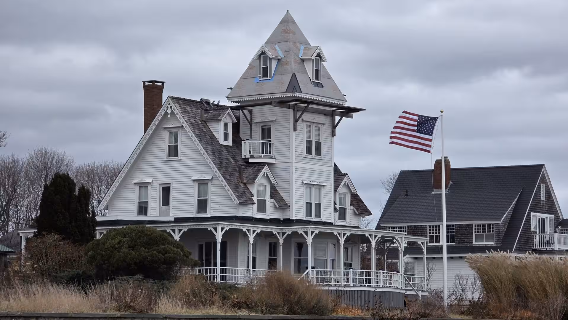 Large Victorian-style coastal house with tower and American flag under dramatic gray skies on the New England shoreline.