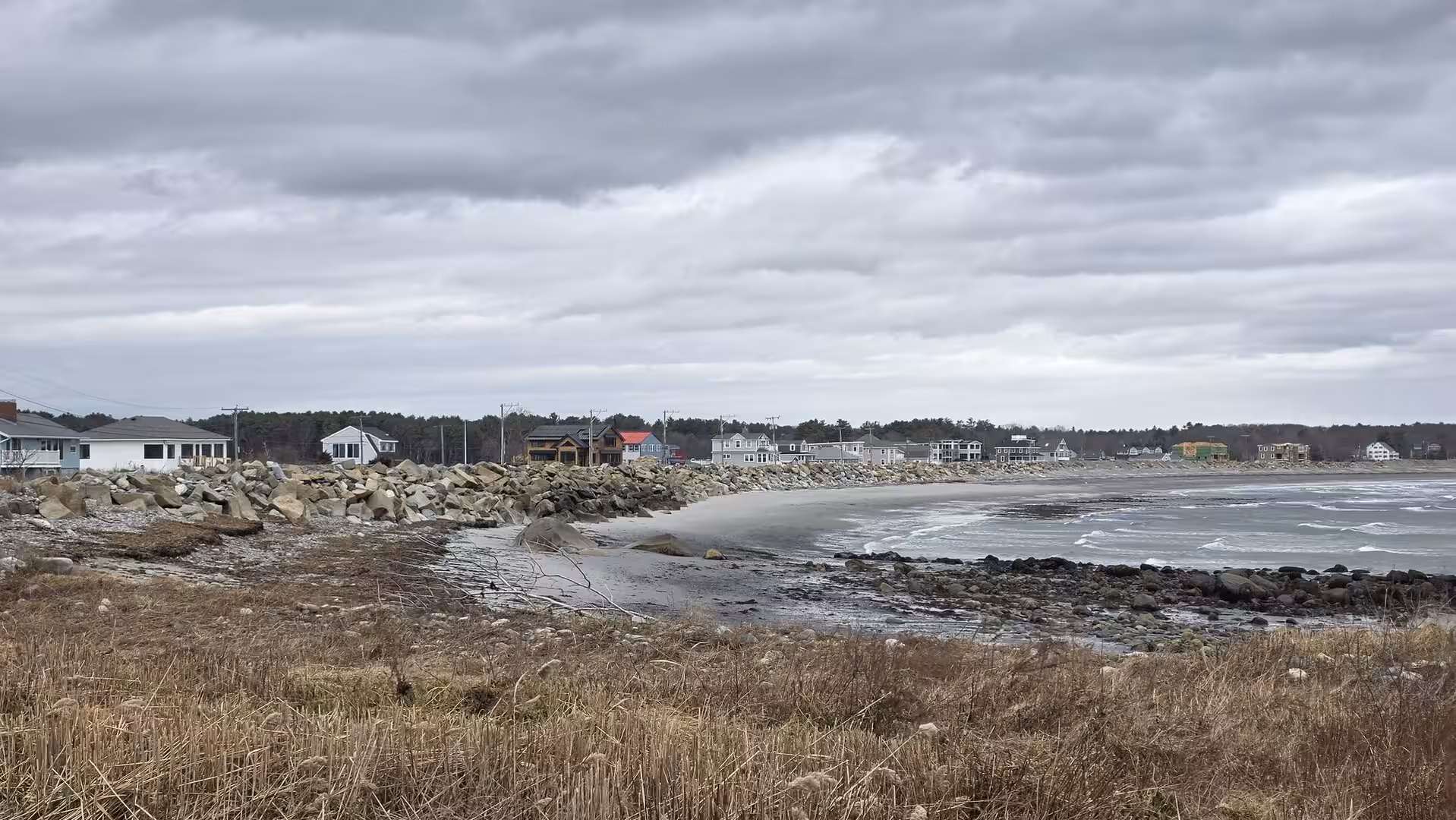 Rocky New England coastline with scattered seaside homes, rough Atlantic surf, and dramatic overcast skies.