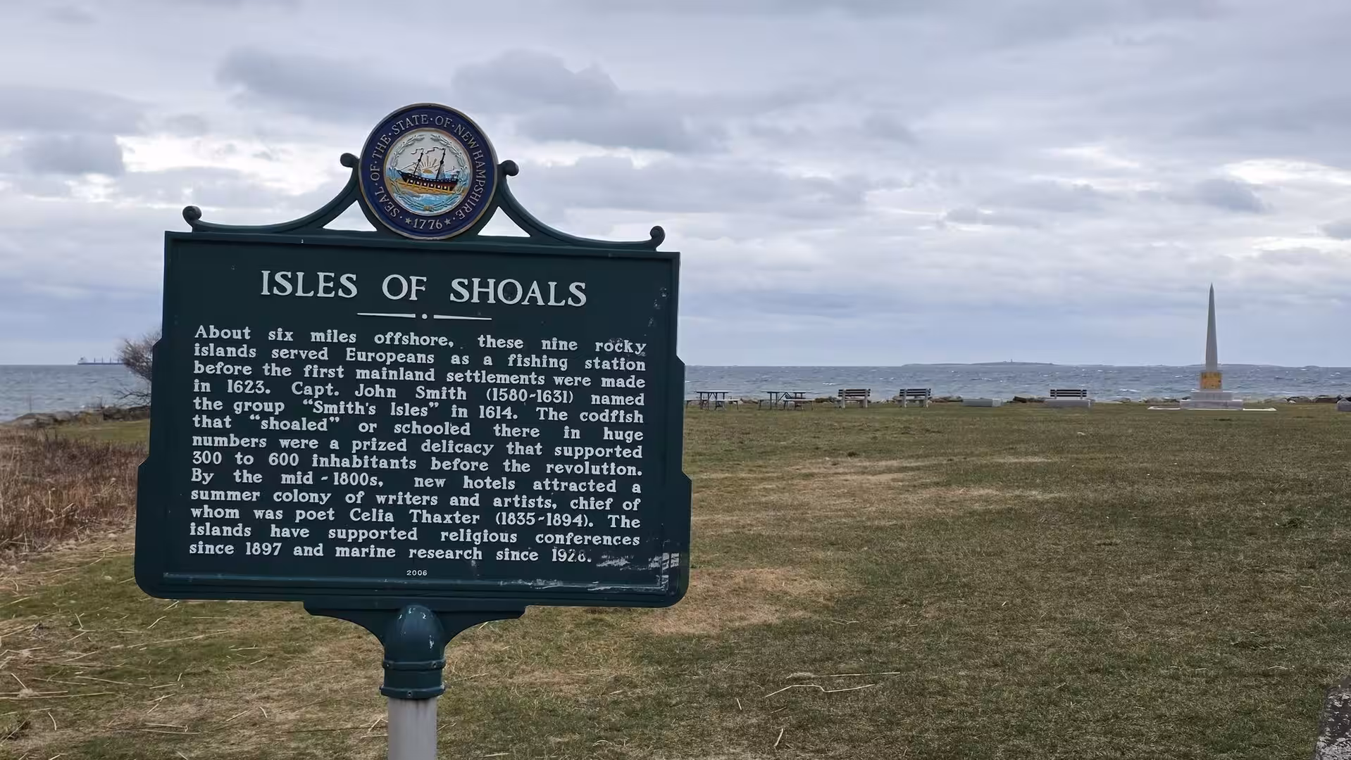 Historic Isles of Shoals information sign beside the Atlantic shoreline with monument and cloudy coastal sky in the background.