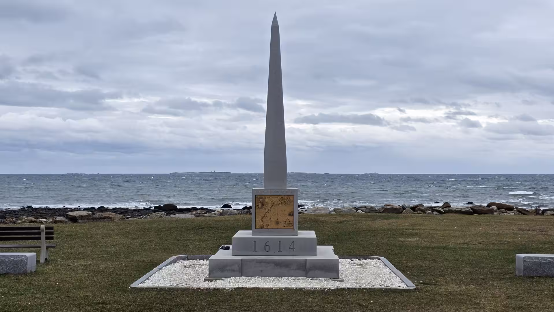 Tall stone obelisk monument marked “1614” standing beside the Atlantic coastline under a cloudy New England sky.