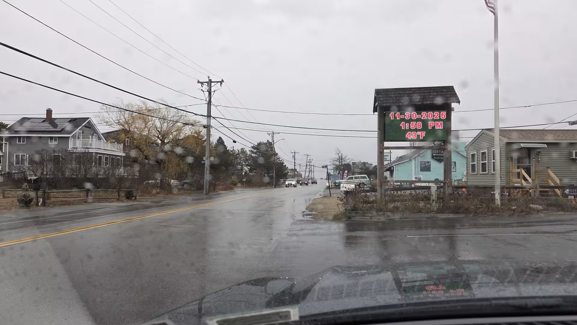 Rainy coastal roadside scene with wet pavement, utility poles, small seaside houses, and an electronic sign displaying date, time, and temperature.