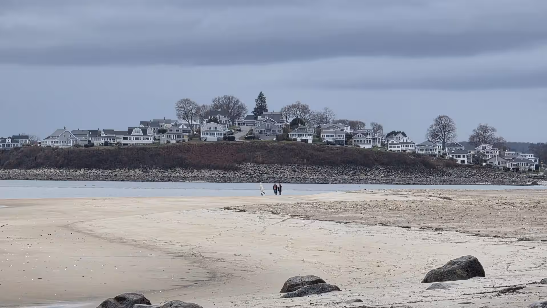 Wide sandy tidal beach with distant coastal homes on a rocky shoreline beneath dark overcast winter clouds.