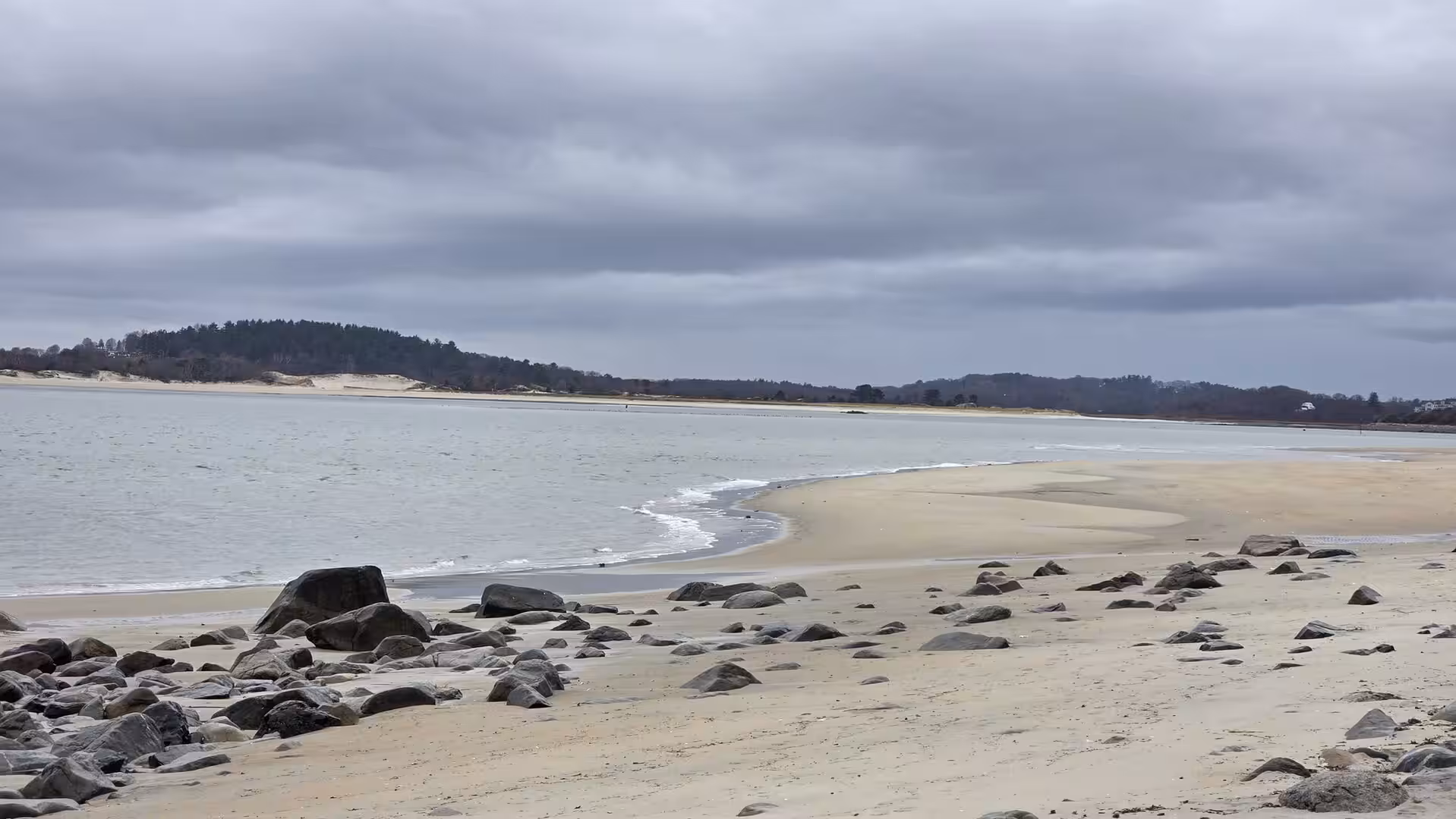 Quiet sandy beach with scattered rocks, calm tidal water, and dramatic grey clouds stretching across a cold New England shoreline.