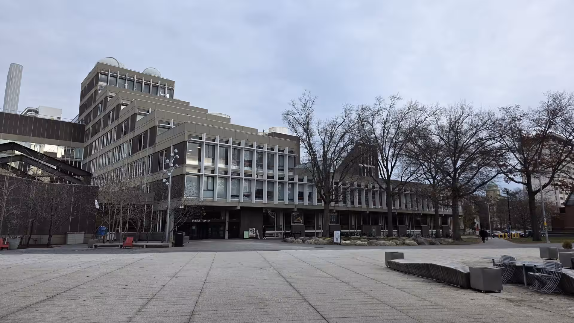 Modern university or research building with glass facades and open plaza surrounded by leafless trees under an overcast winter sky.