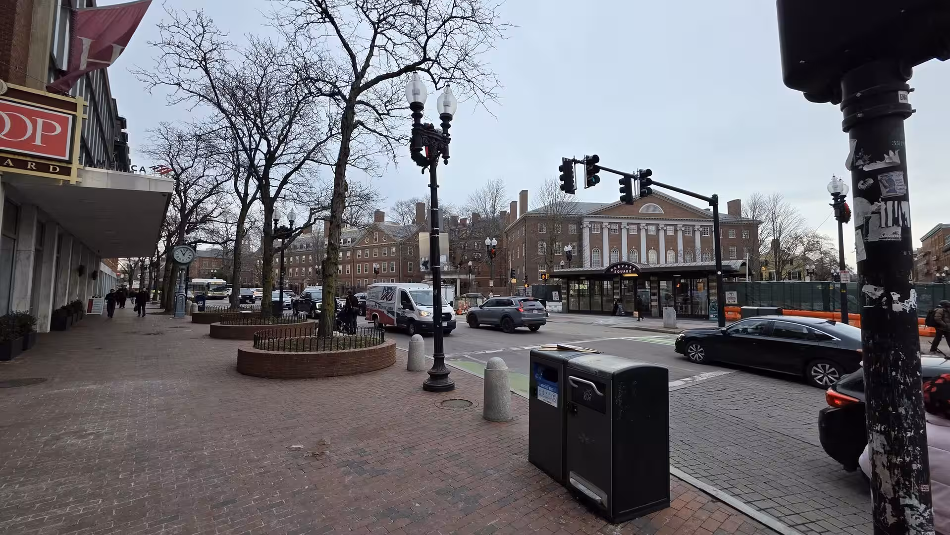 Street scene near Harvard Square with historic brick buildings, leafless trees, traffic lights, and pedestrians on a cloudy winter day.