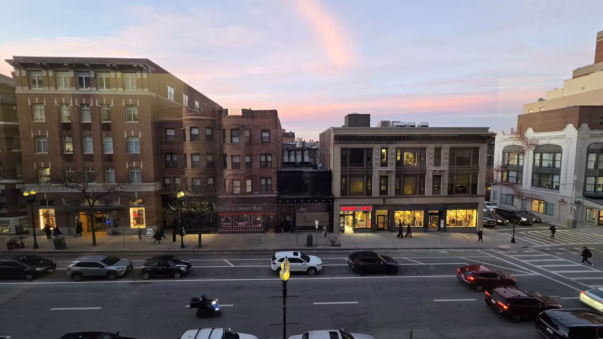 Evening street scene in Boston with historic brick buildings, illuminated storefronts, moving traffic, and pastel sunset clouds overhead.