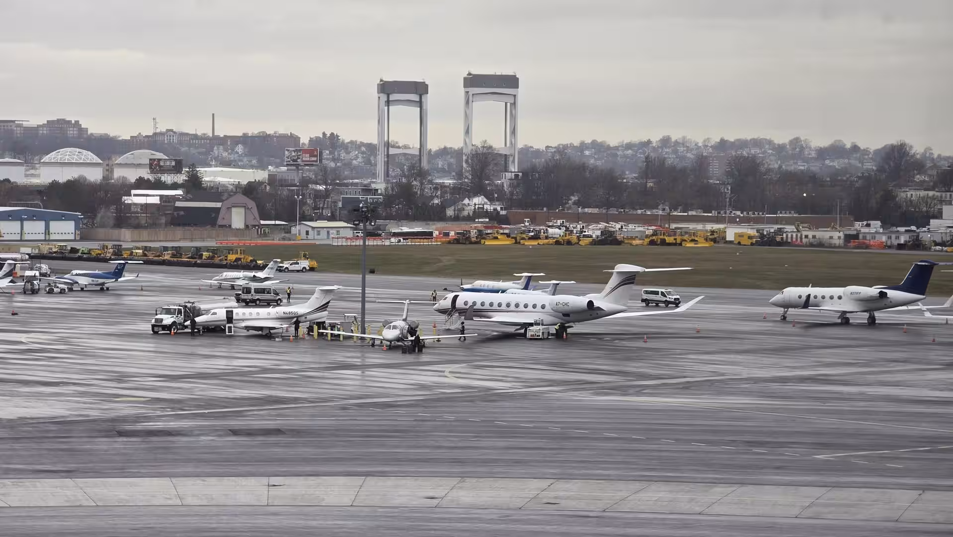 Private business jets parked on a rain-soaked airport apron under overcast skies with airport infrastructure and city skyline in the distance.