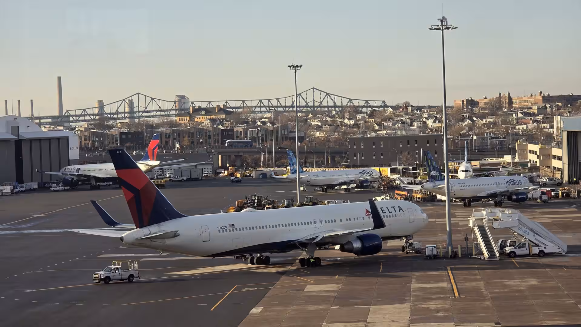 Delta Air Lines aircraft parked at an airport apron with JetBlue planes and an urban skyline featuring a large steel bridge in the background.