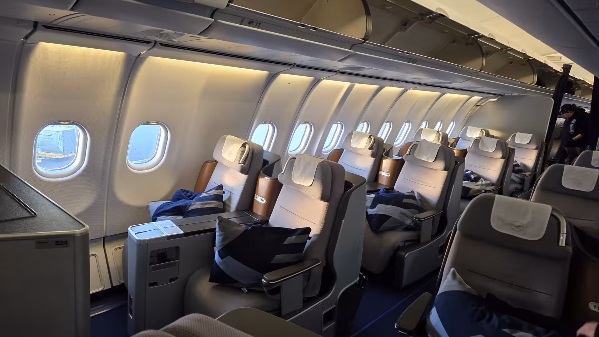 Modern premium economy or business class cabin interior on a Lufthansa widebody aircraft with spacious grey seats, large windows, and warm ambient lighting during boarding.
