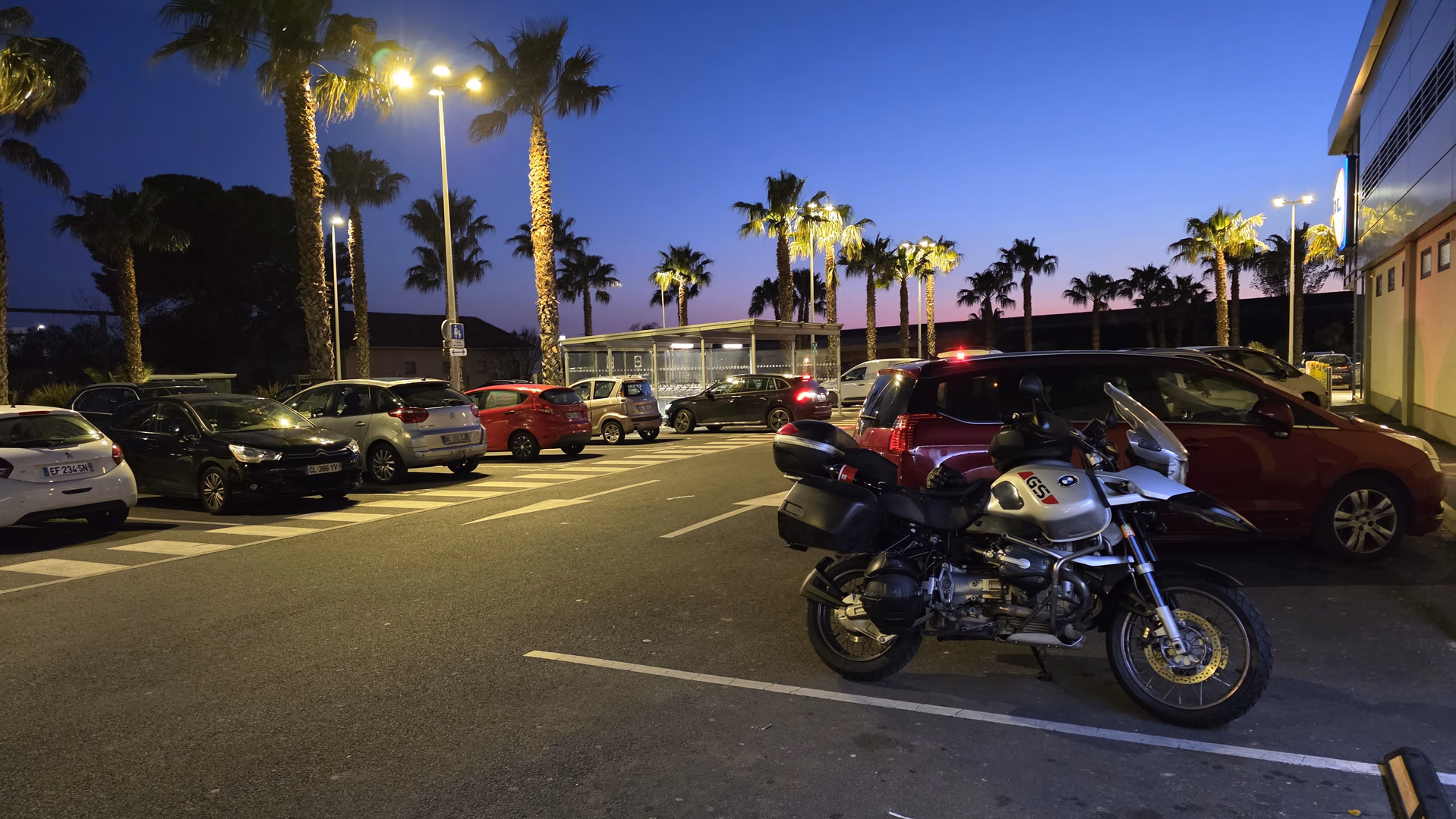 Touring motorcycle parked in a palm-lined parking area at dusk with illuminated streetlights and a colorful evening sky.