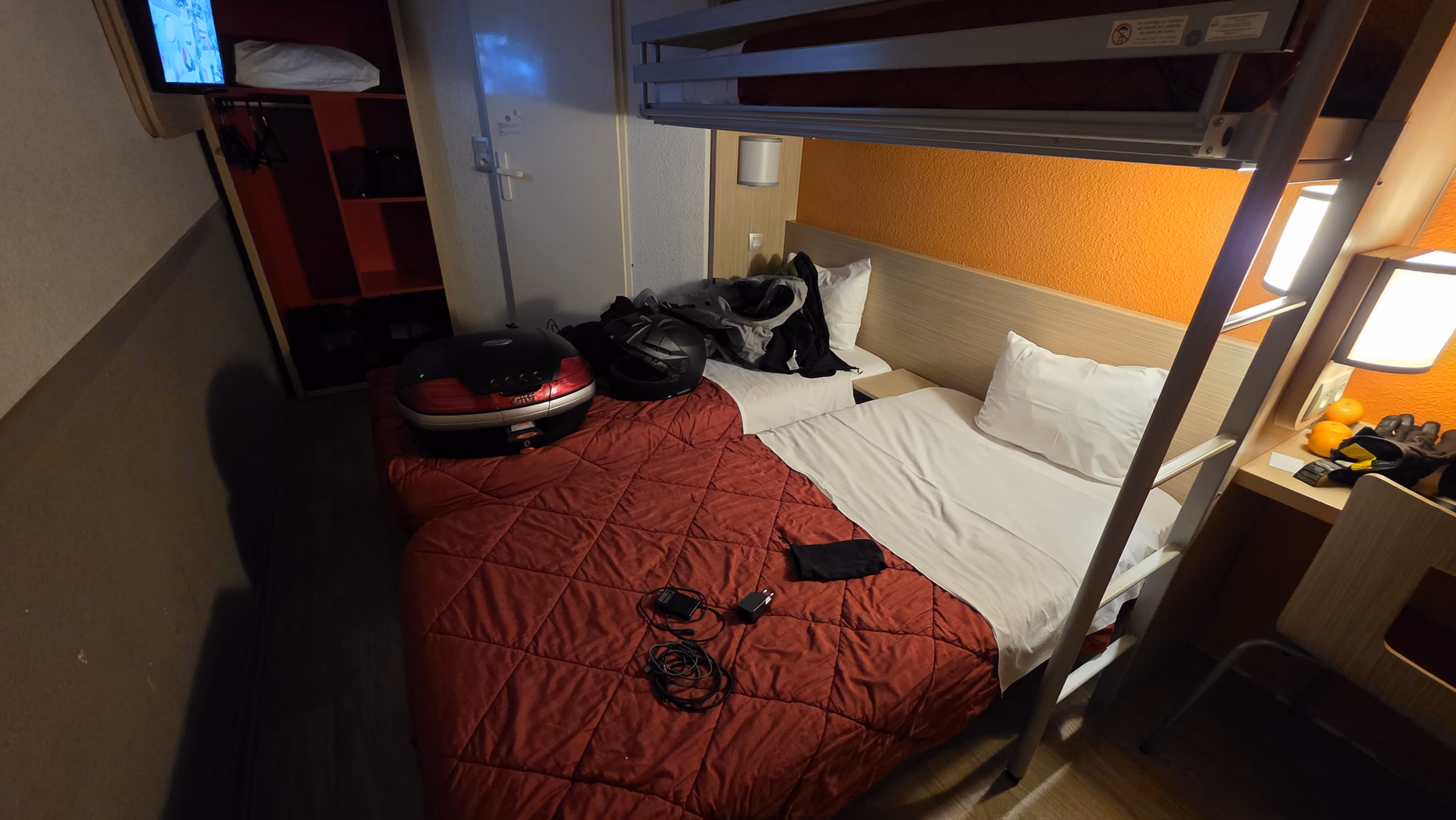 Compact hotel room with a double bed, upper bunk bed, desk, and travel gear.