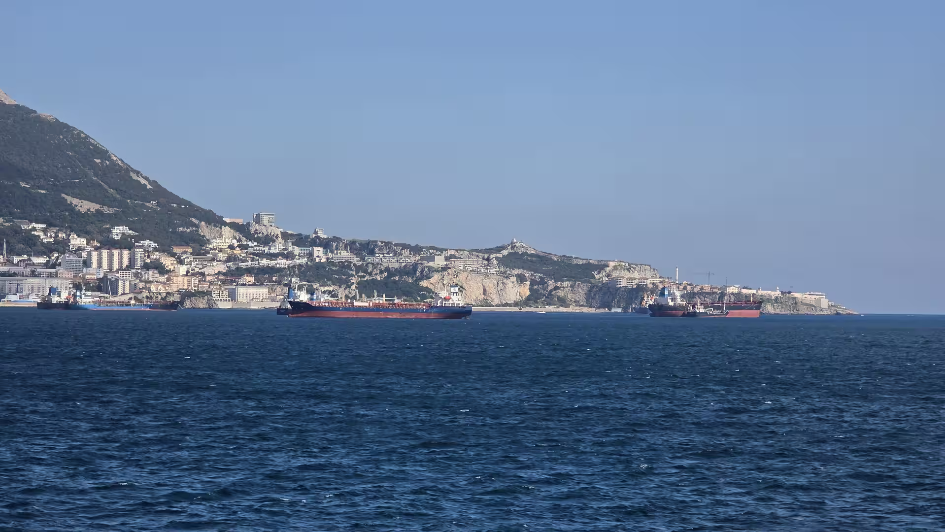 Cargo tankers anchored offshore near Gibraltar with the coastline and cliffs in the background.