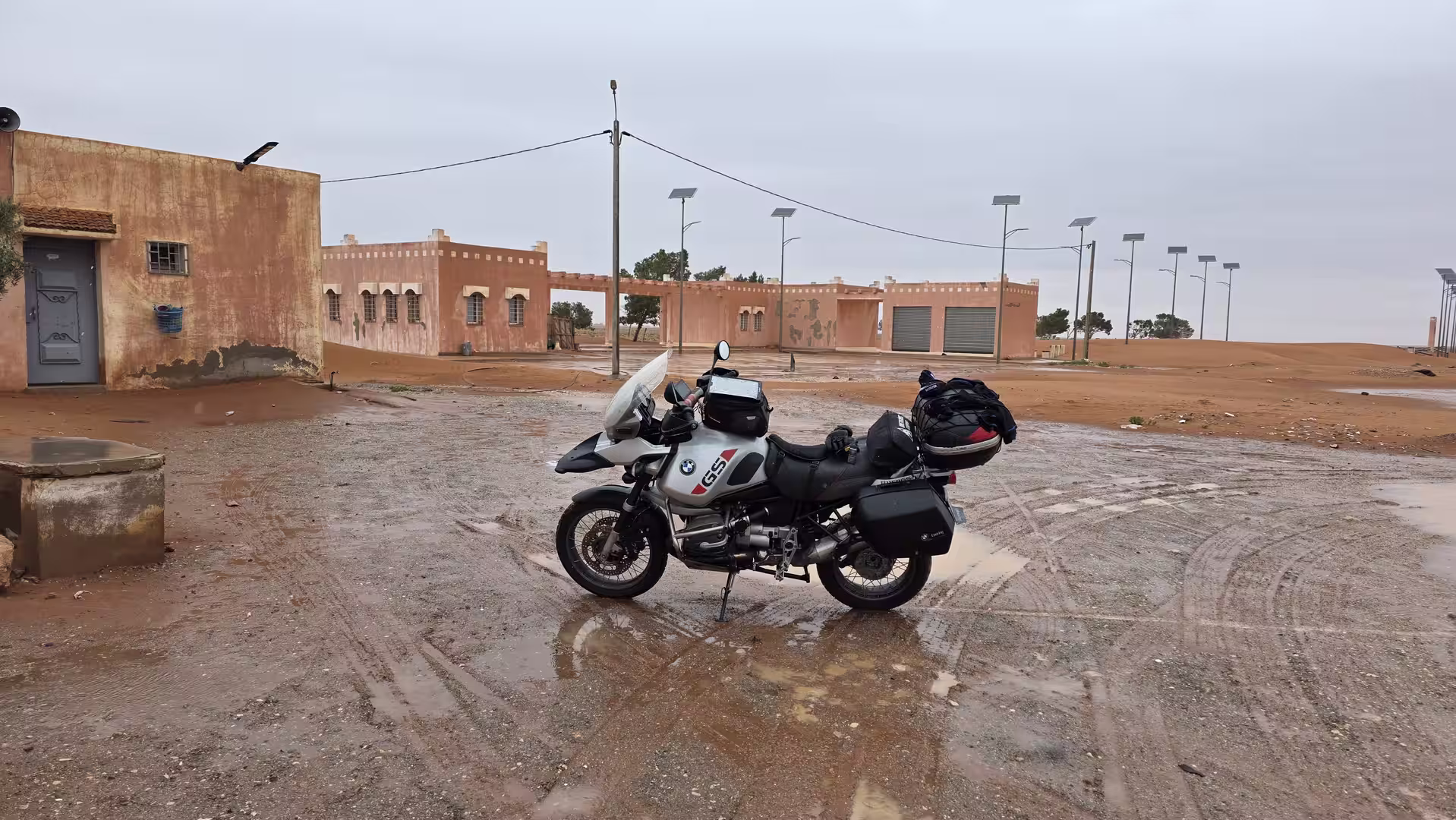 A BMW adventure motorcycle parked on a wet desert square under an overcast sky.
