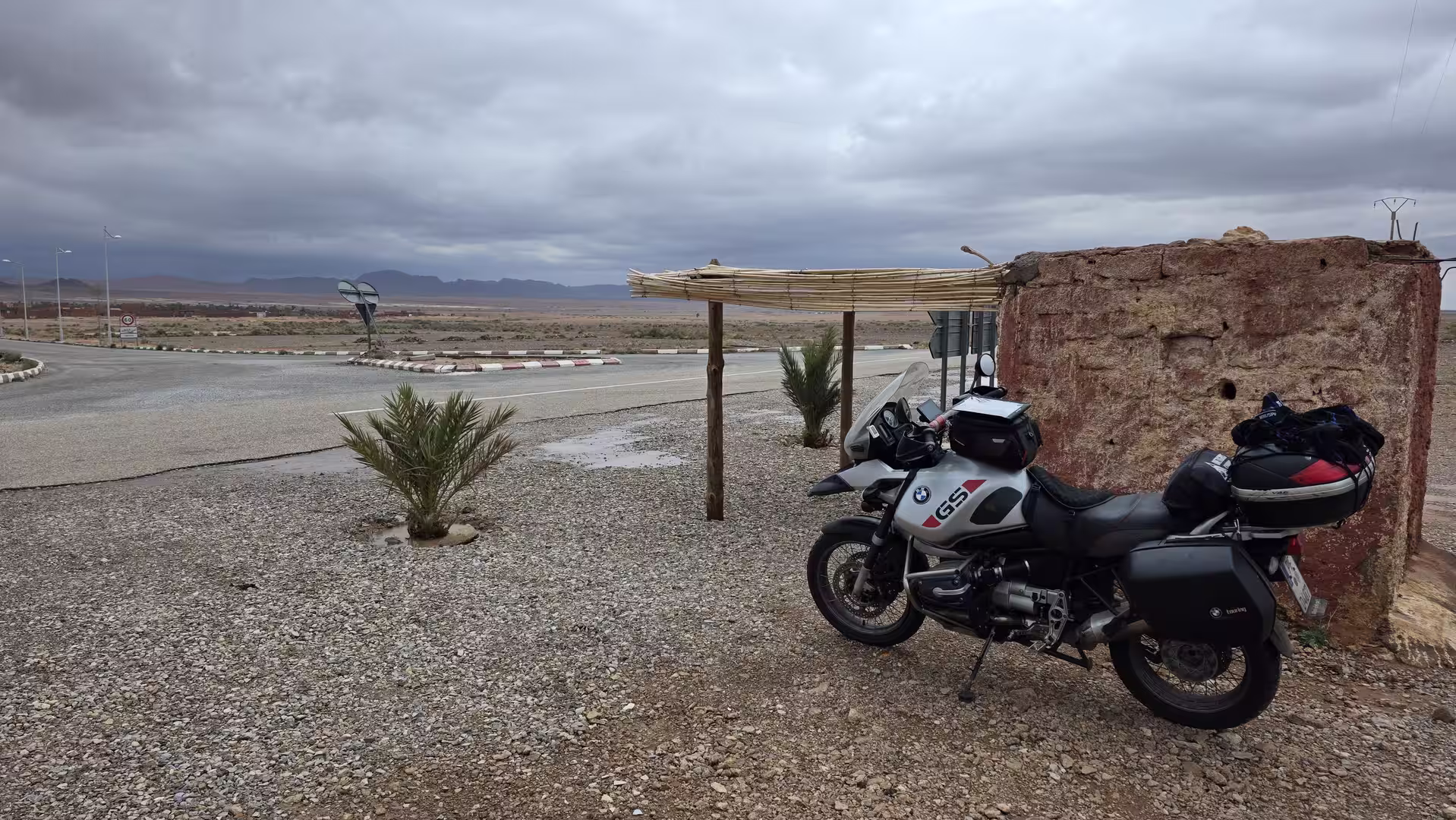 A BMW GS motorcycle parked beside a roadside shelter beneath dark storm clouds in eastern Morocco.