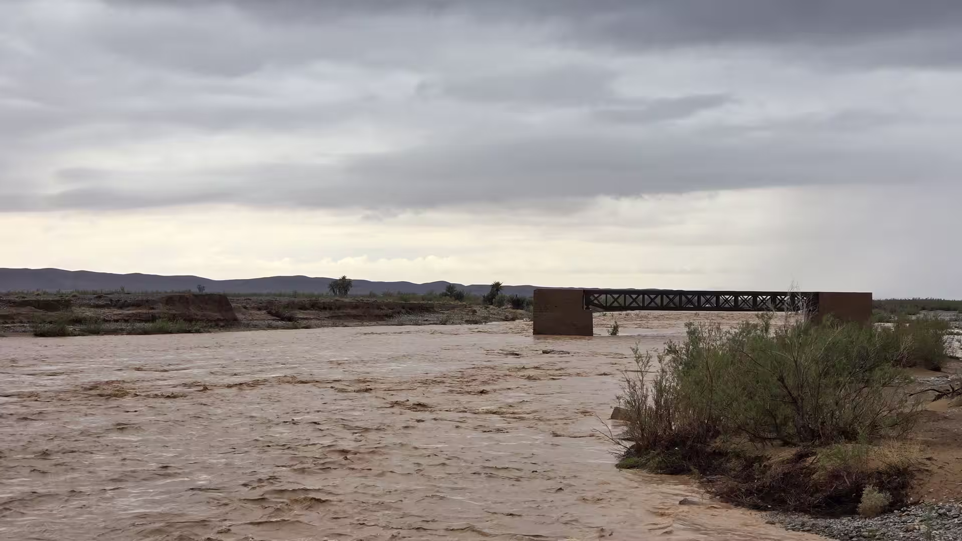 A swollen desert river carrying muddy floodwaters beneath dark storm clouds in eastern Morocco.