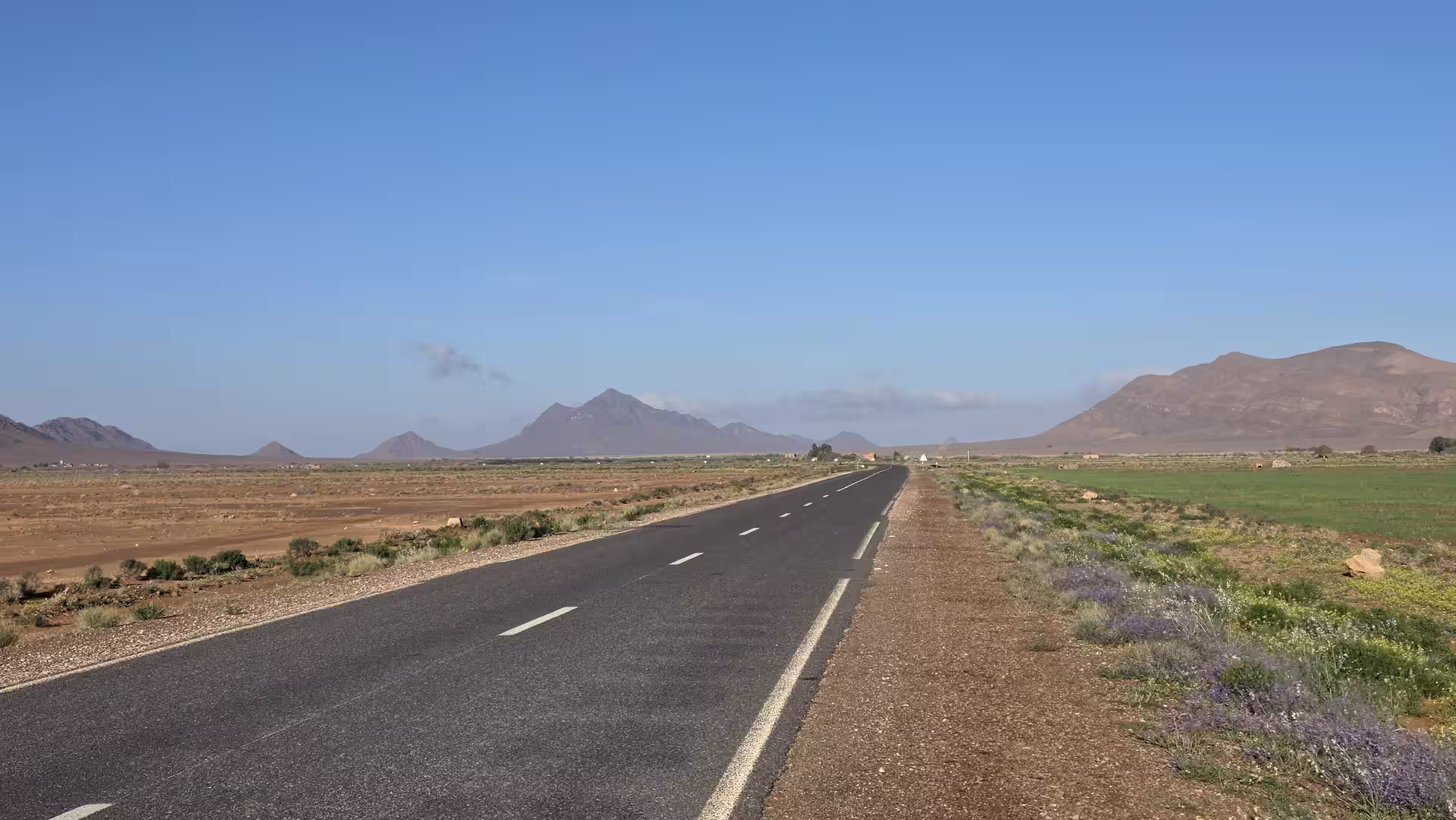 Empty road crossing a broad valley with mountain ranges in eastern Morocco under a clear blue sky.