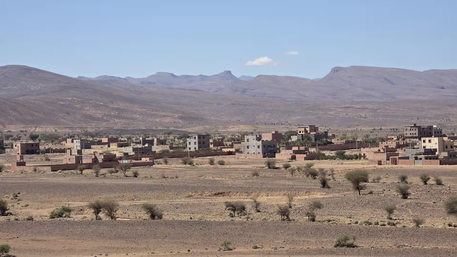 Small settlement with scattered buildings in an arid plain beneath layered mountain ridges in eastern Morocco.