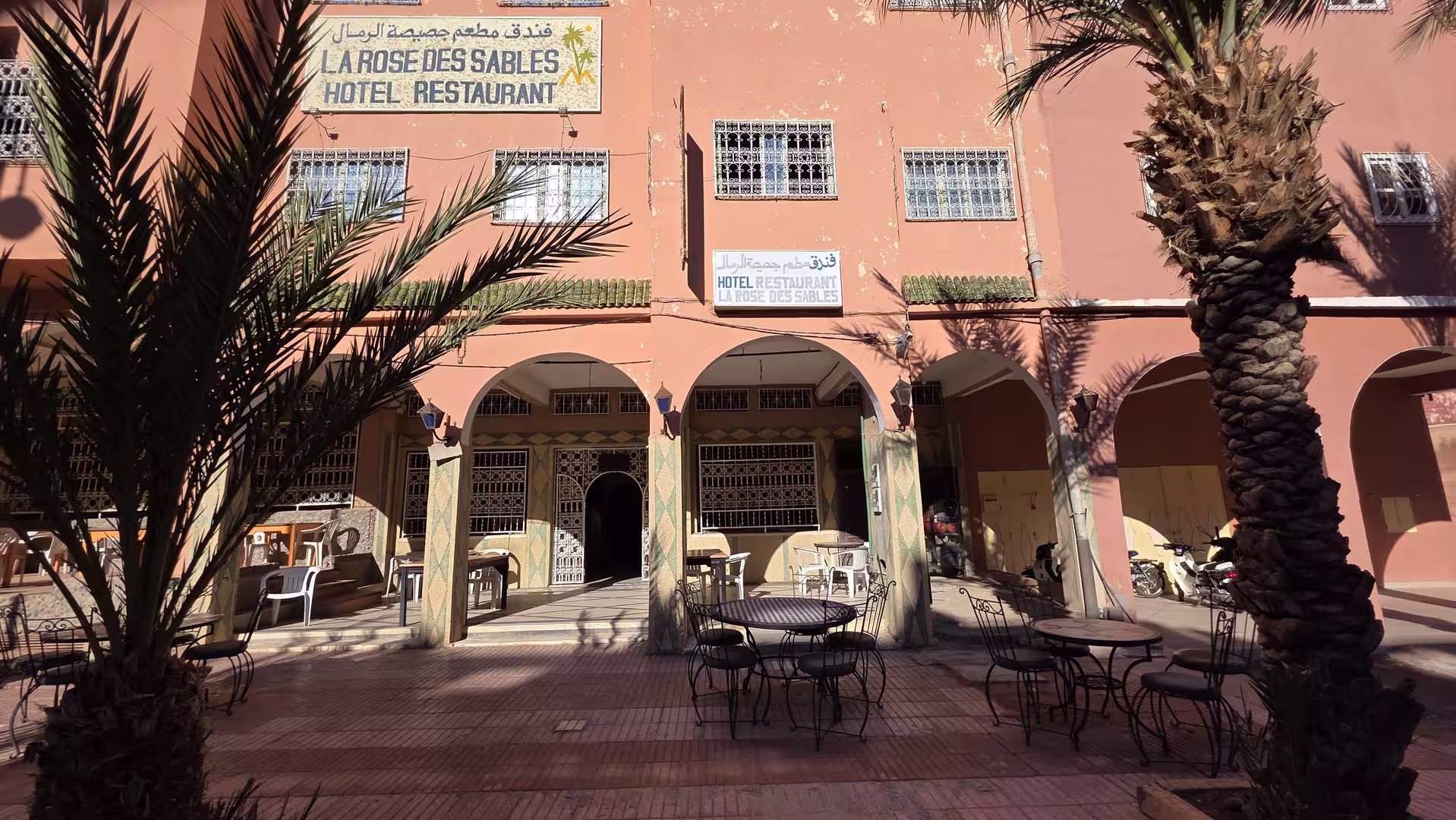 Courtyard entrance of Hotel La Rose des Sables with palm trees, arcades, and outdoor seating in Zagora, Morocco.
