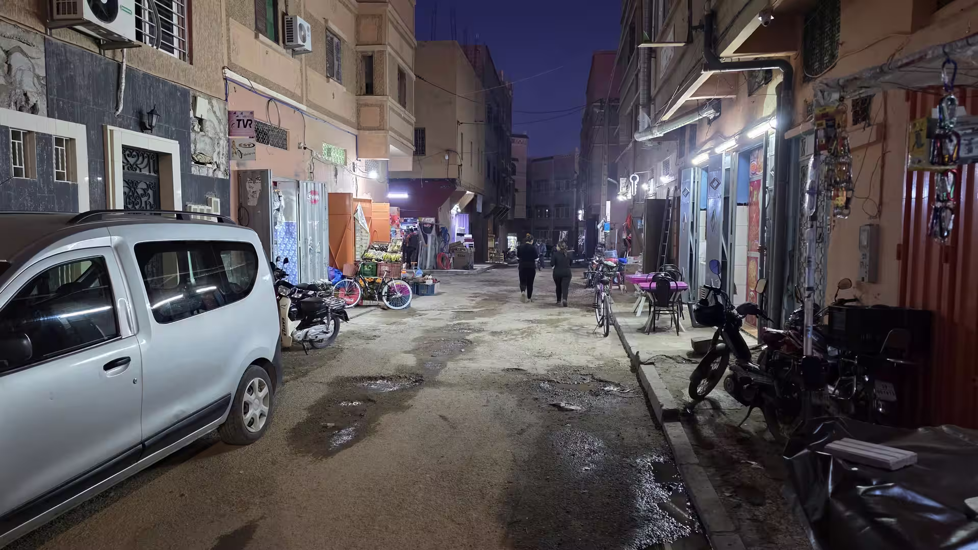 Narrow street in Zagora at dusk with parked vehicles, small shops, bicycles, and people walking under bright storefront lights.