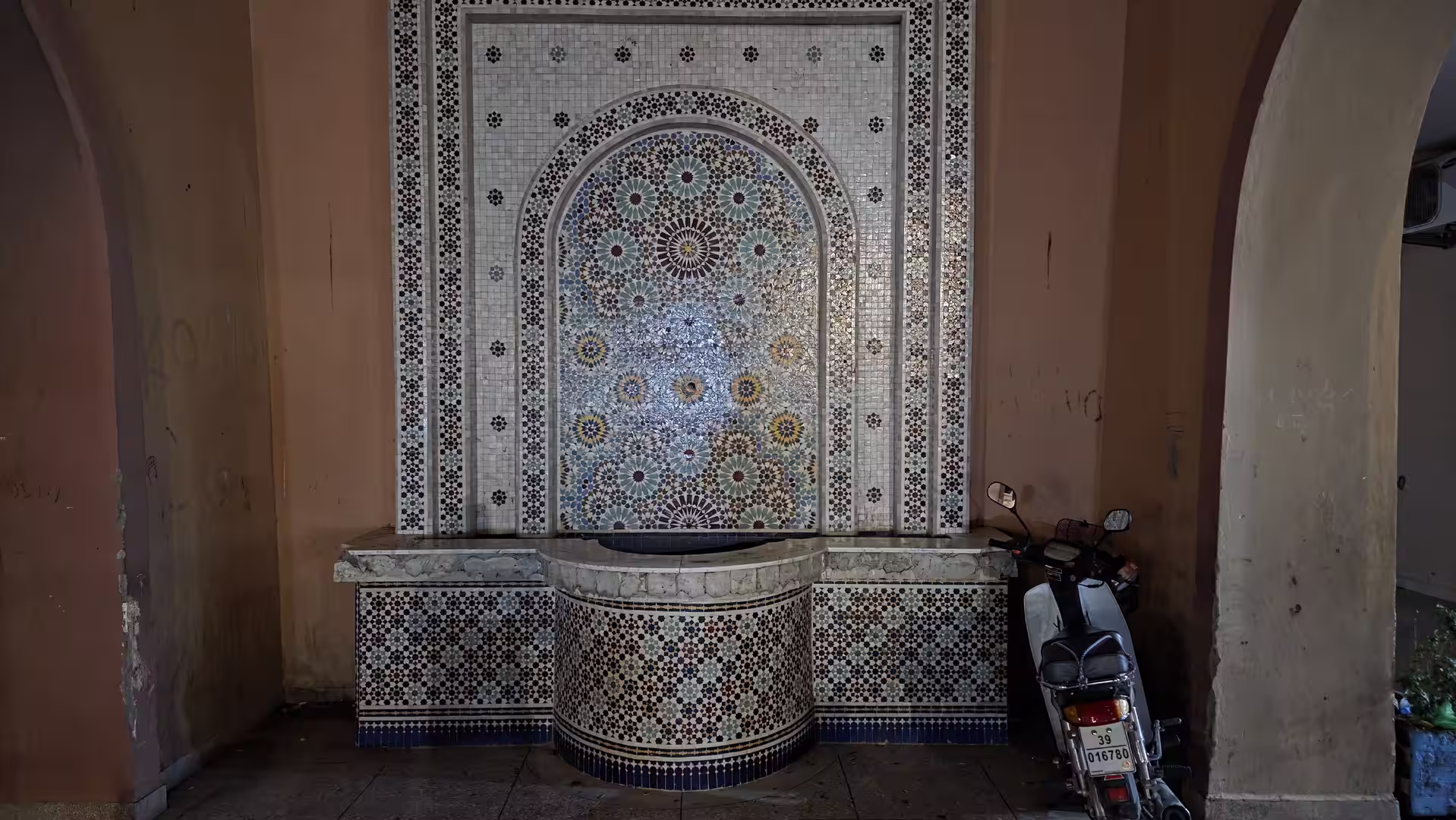 Decorative tiled fountain set inside an arched alcove in Zagora with intricate geometric mosaic patterns and a parked scooter nearby.