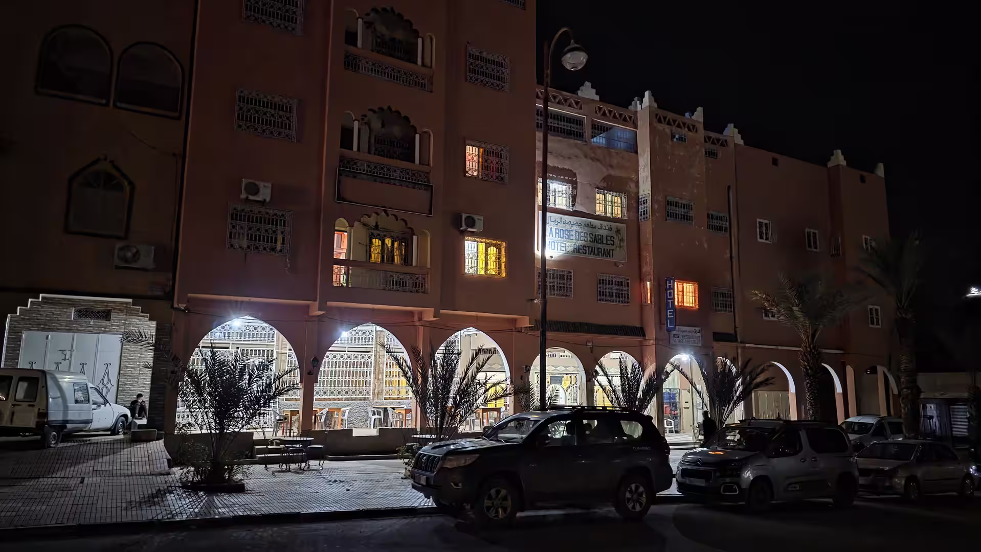 Night view of La Rose des Sables hotel in Zagora with illuminated arcades, parked cars, and warm colored façade.