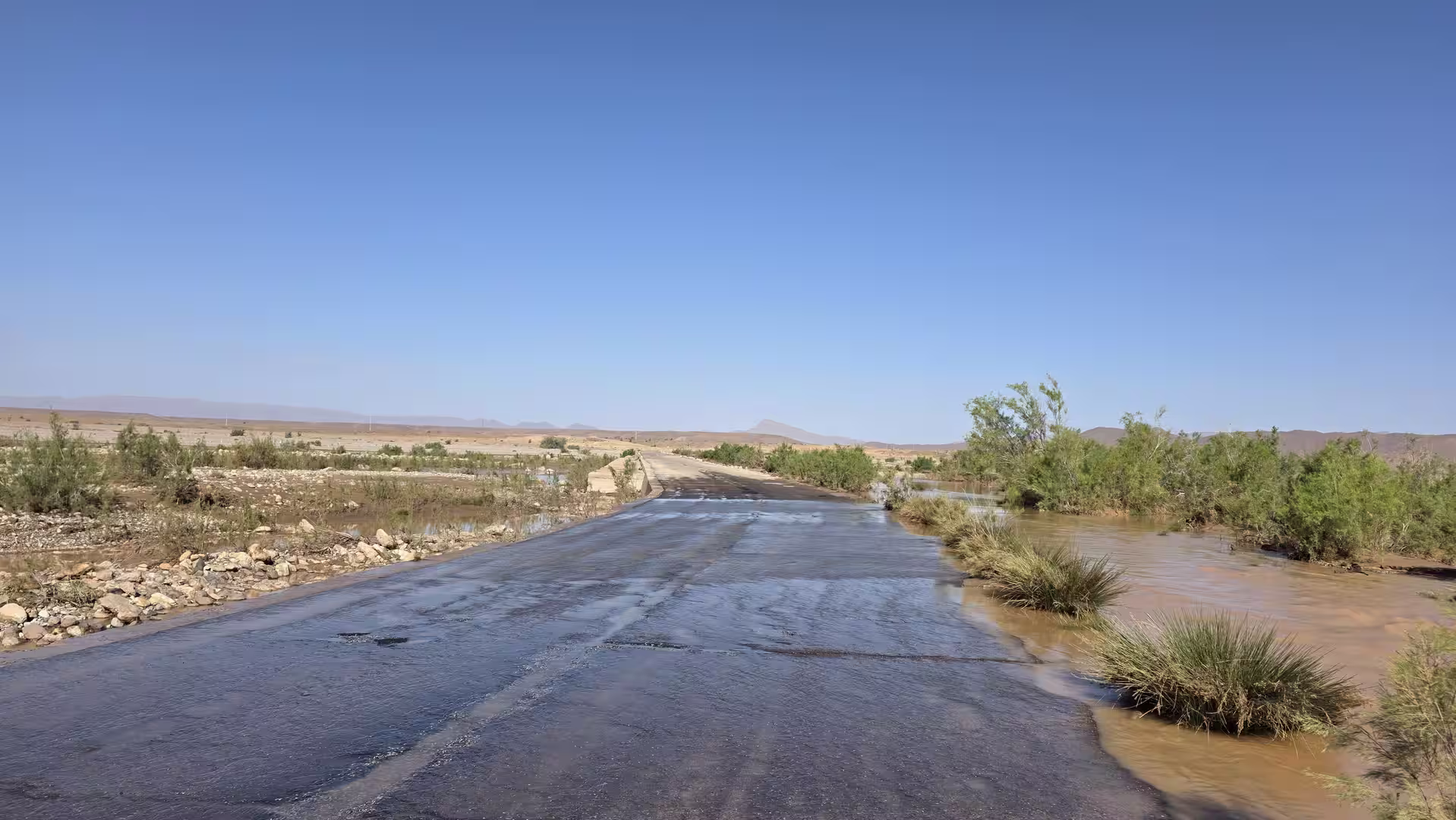 Road crossing partially covered by shallow floodwater in an arid landscape with sparse vegetation and clear blue sky.