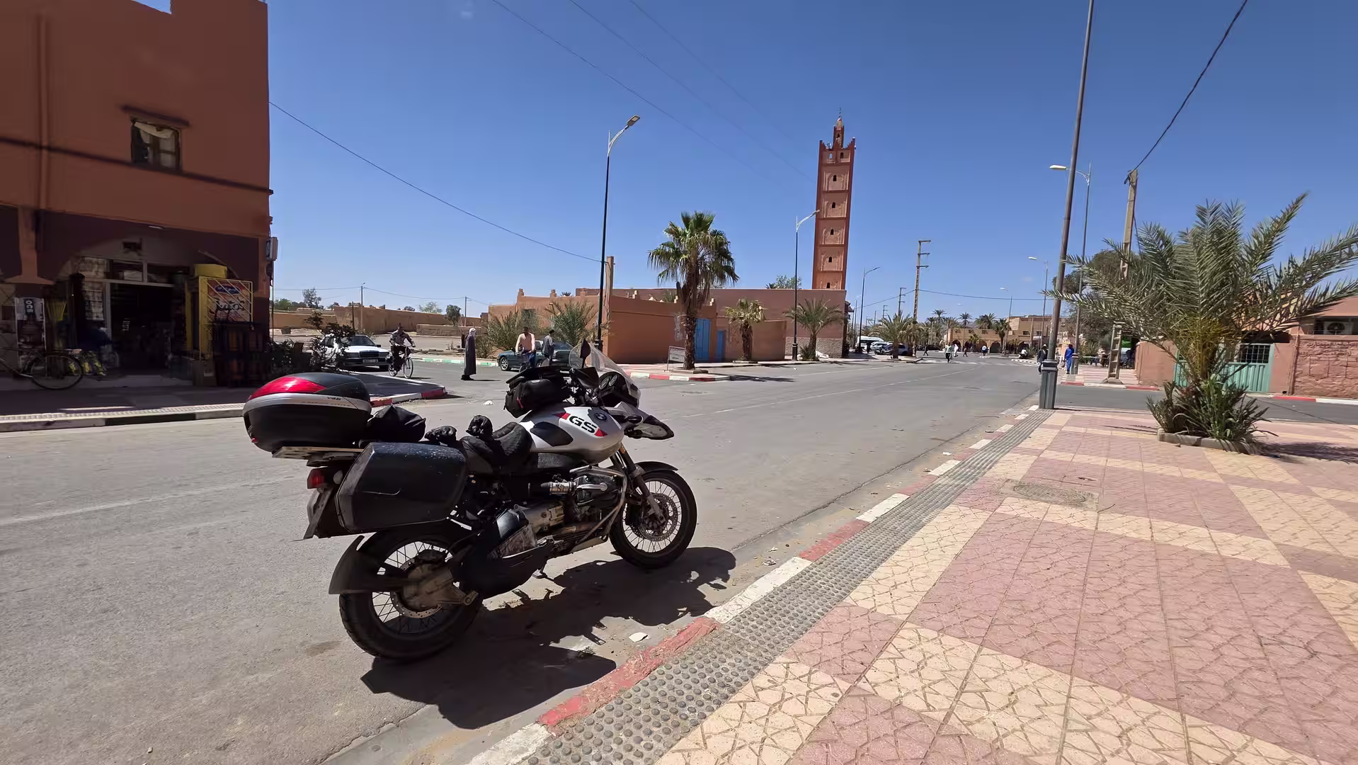 Touring motorcycle parked along a wide street lined with palm trees and low-rise buildings, with a tall mosque minaret in the background.