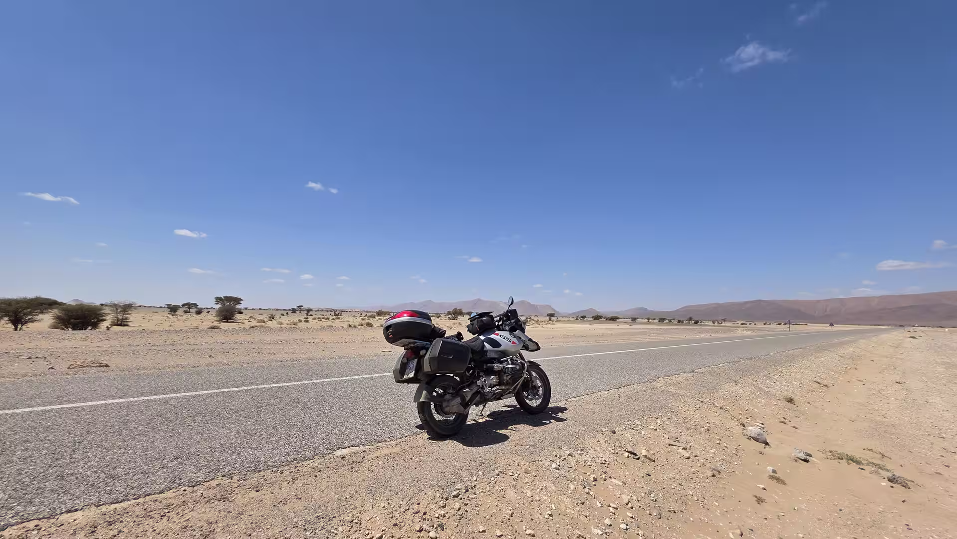 Touring motorcycle parked beside a long paved road crossing an open desert plain with distant mountains under a clear blue sky.