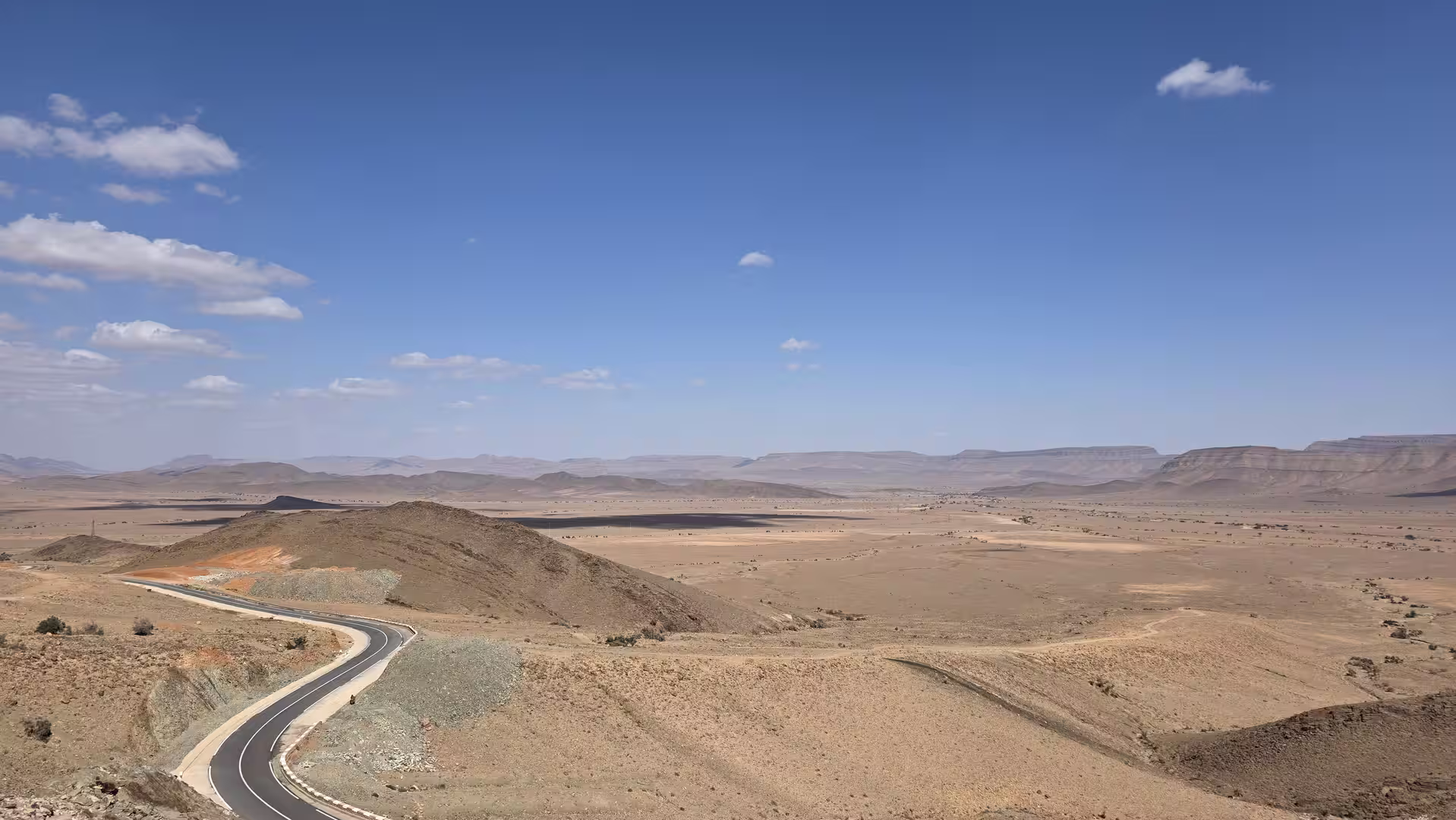 Wide desert landscape with a curving paved road descending through rolling hills toward distant plateaus under a blue sky.