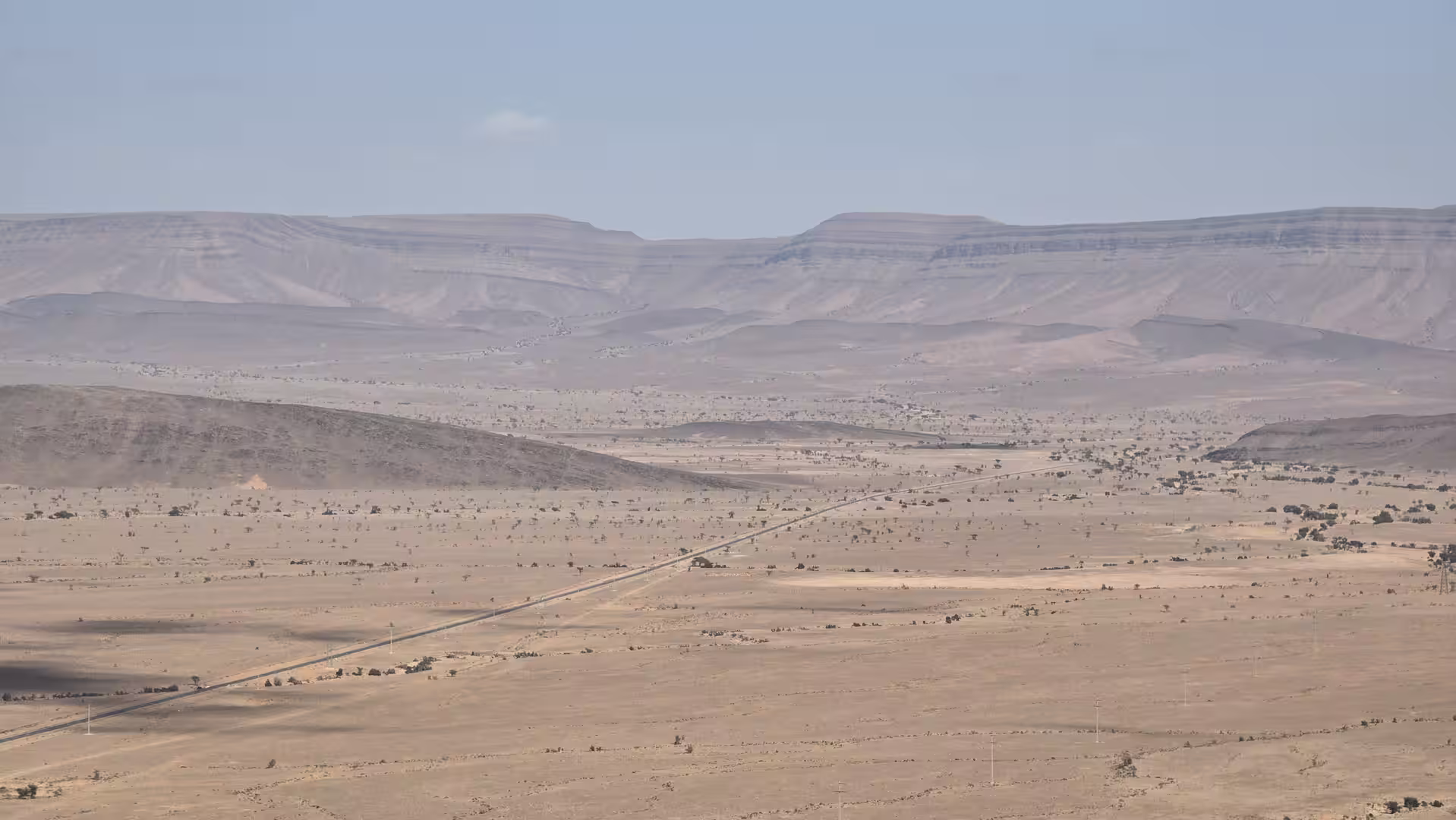 Wide view across an arid valley with a straight road leading toward layered sandstone plateaus in the distance.