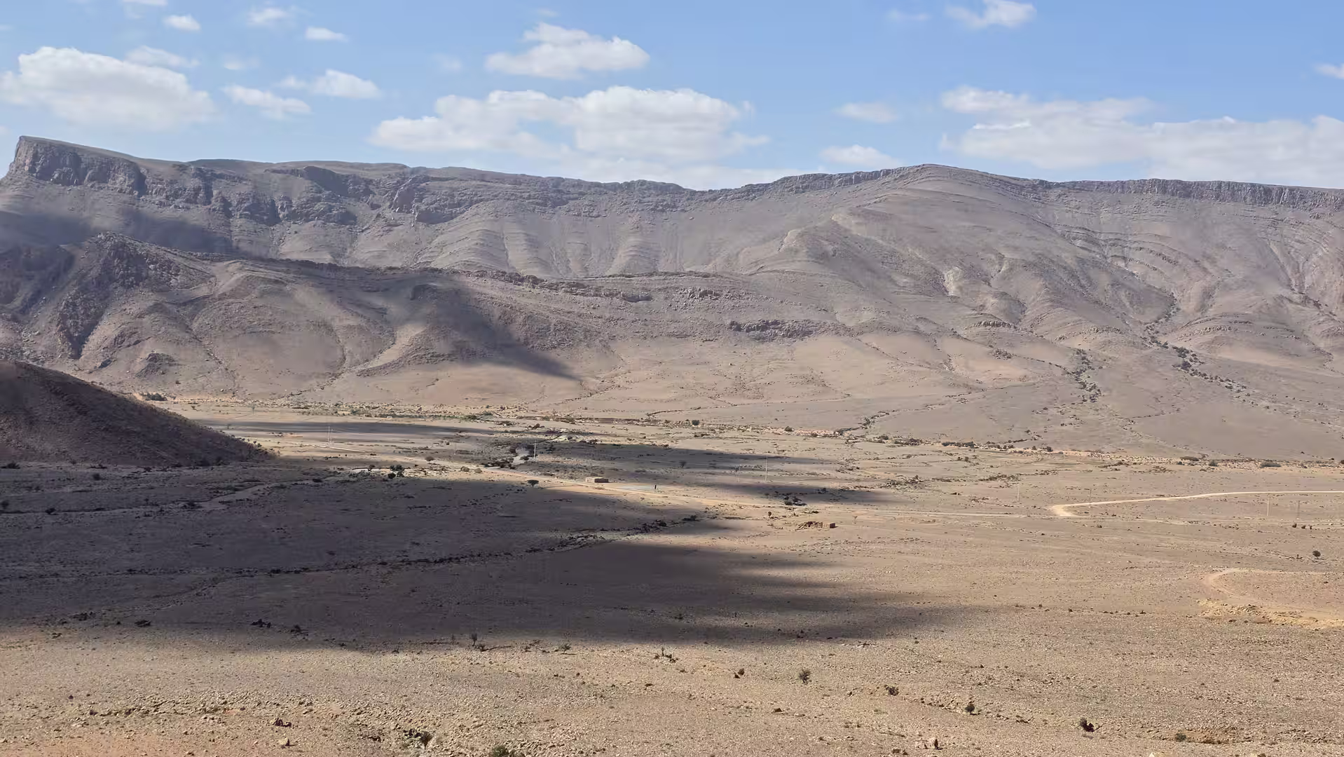 Broad rocky valley surrounded by layered desert mountains with long cloud shadows across the plain.