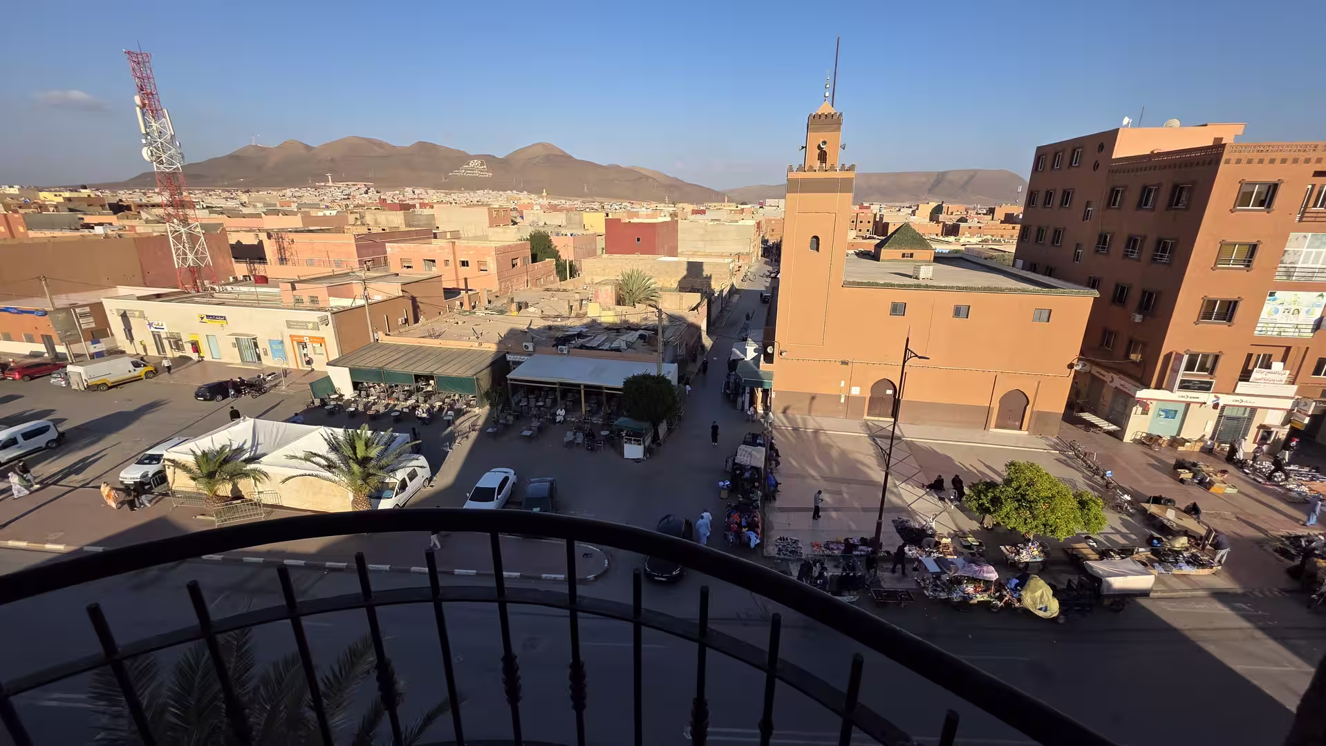 View from a balcony over Guelmim, Morocco, showing a mosque with a tall minaret, surrounding urban streets and the Anti-Atlas mountains in the distance.