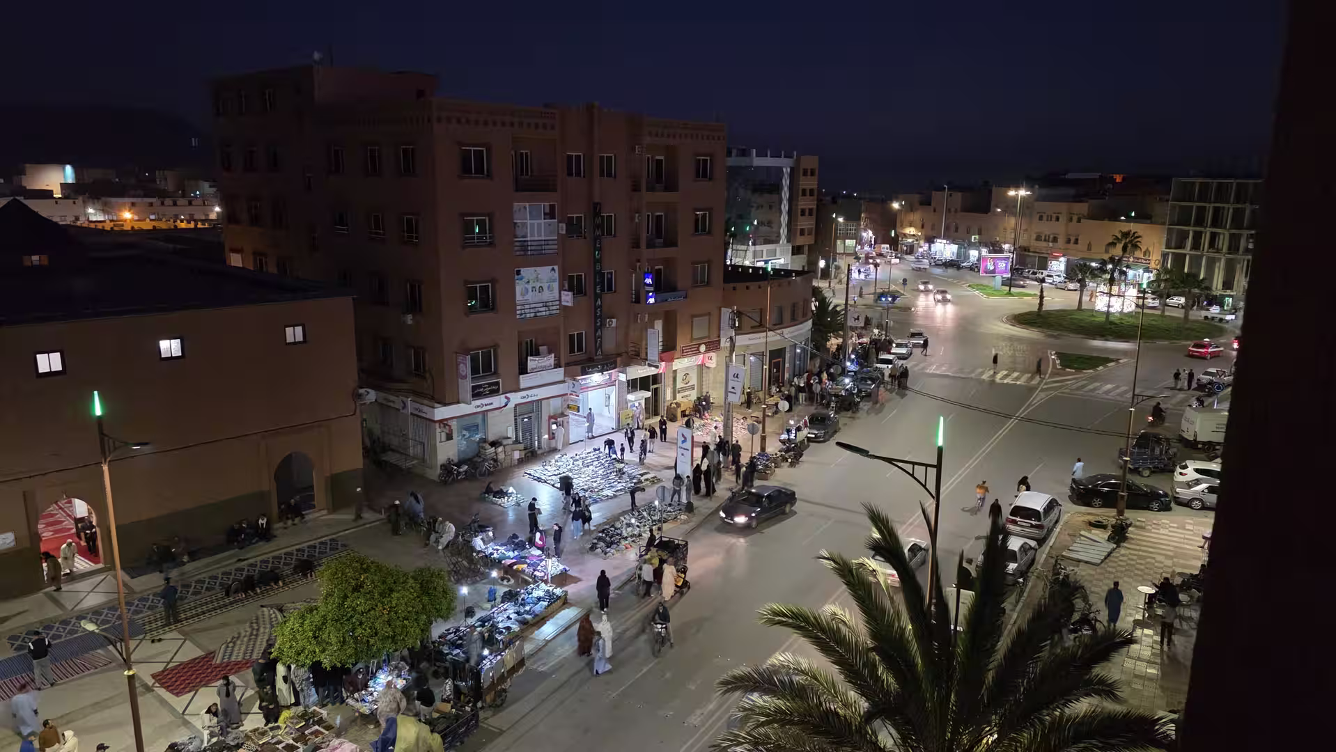 Nighttime view over a lively street intersection in Guelmim, Morocco, with illuminated market stalls, pedestrians, palm trees and city buildings.